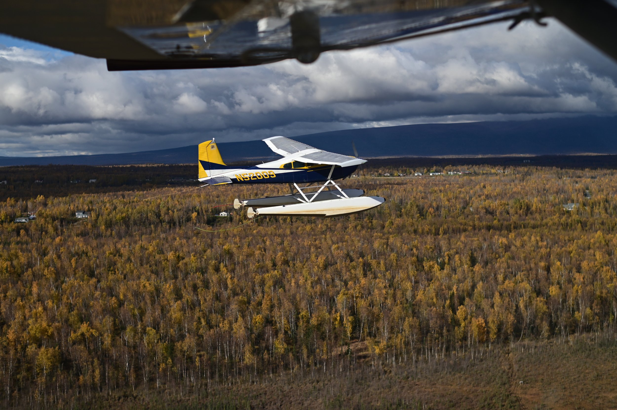 A small seaplane flying over a forest with autumn foliage, seen from another aircraft.