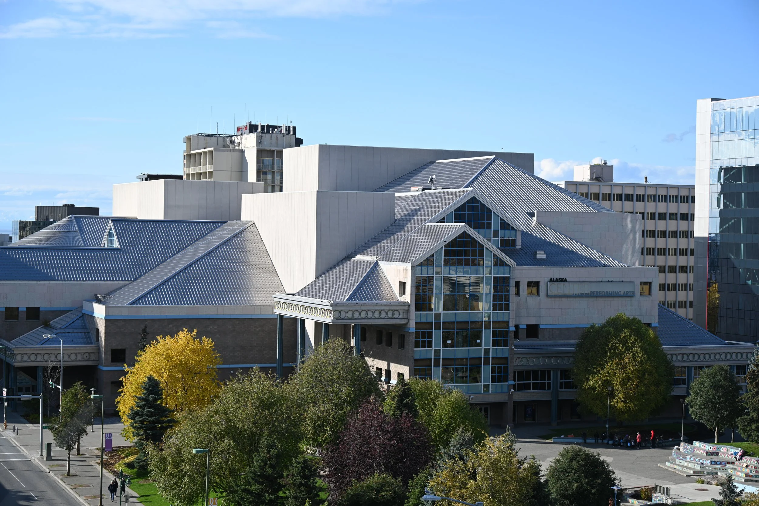 A large modern building with multiple glass windows and metallic roofing, surrounded by trees and a street with pedestrians.