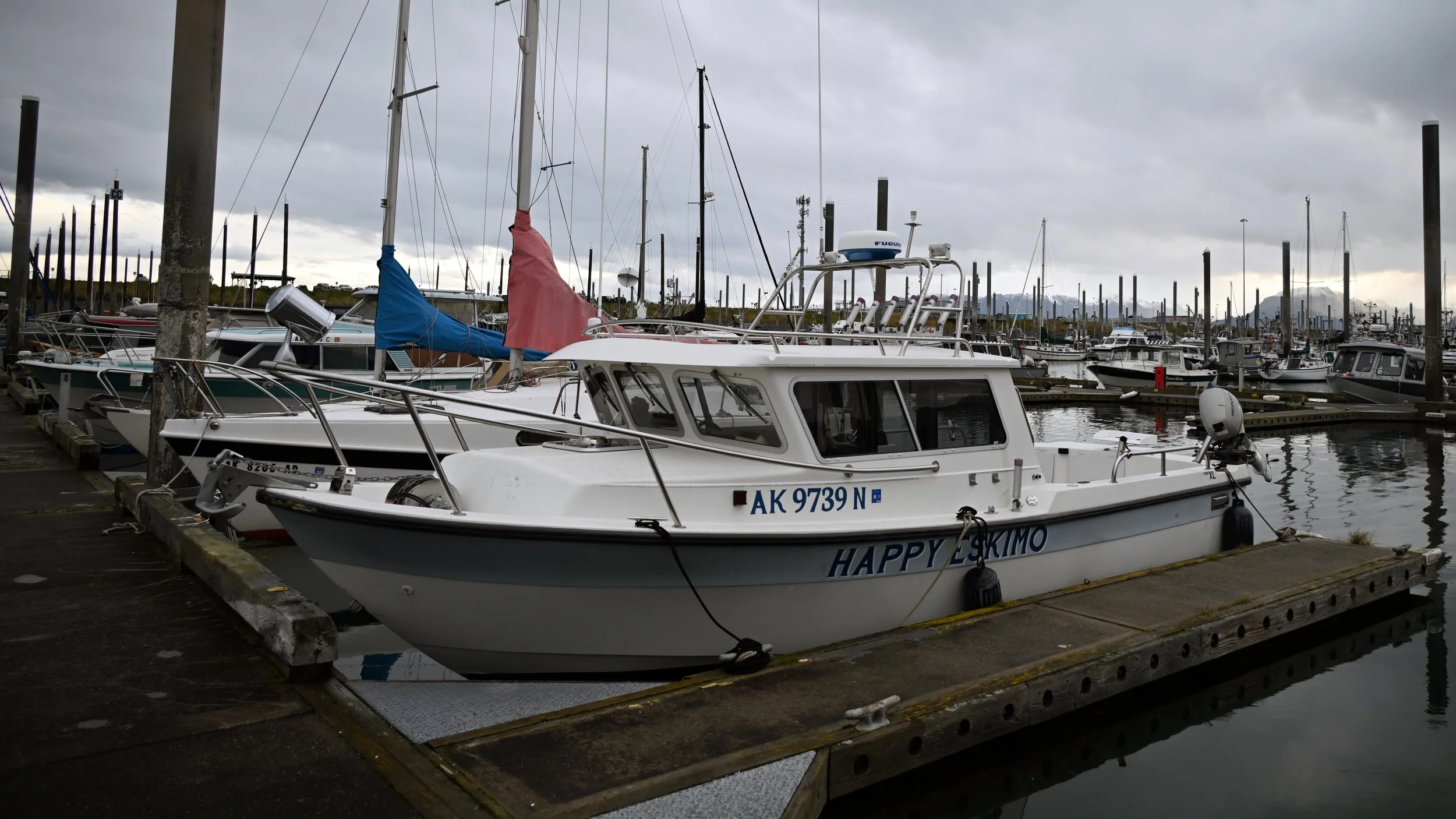 A white boat named 'HAPPY ESKIMO' docked at a marina on a cloudy day, with many other boats and sailboats in the background.