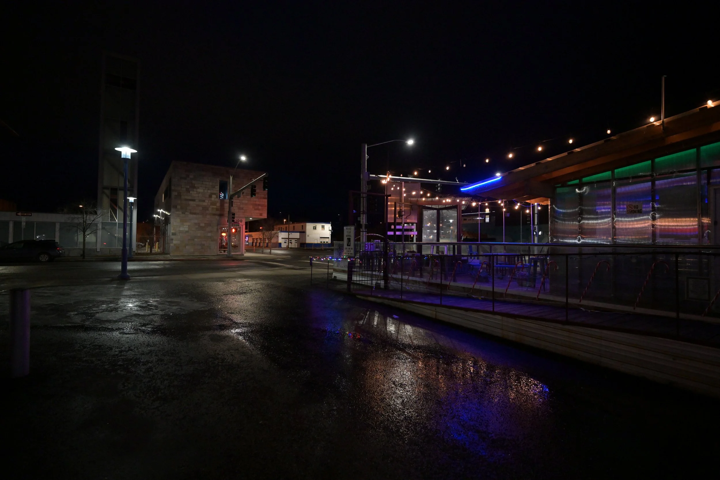 Empty city street at night with wet pavement, illuminated by street lights and colorful neon lights on a building to the right.