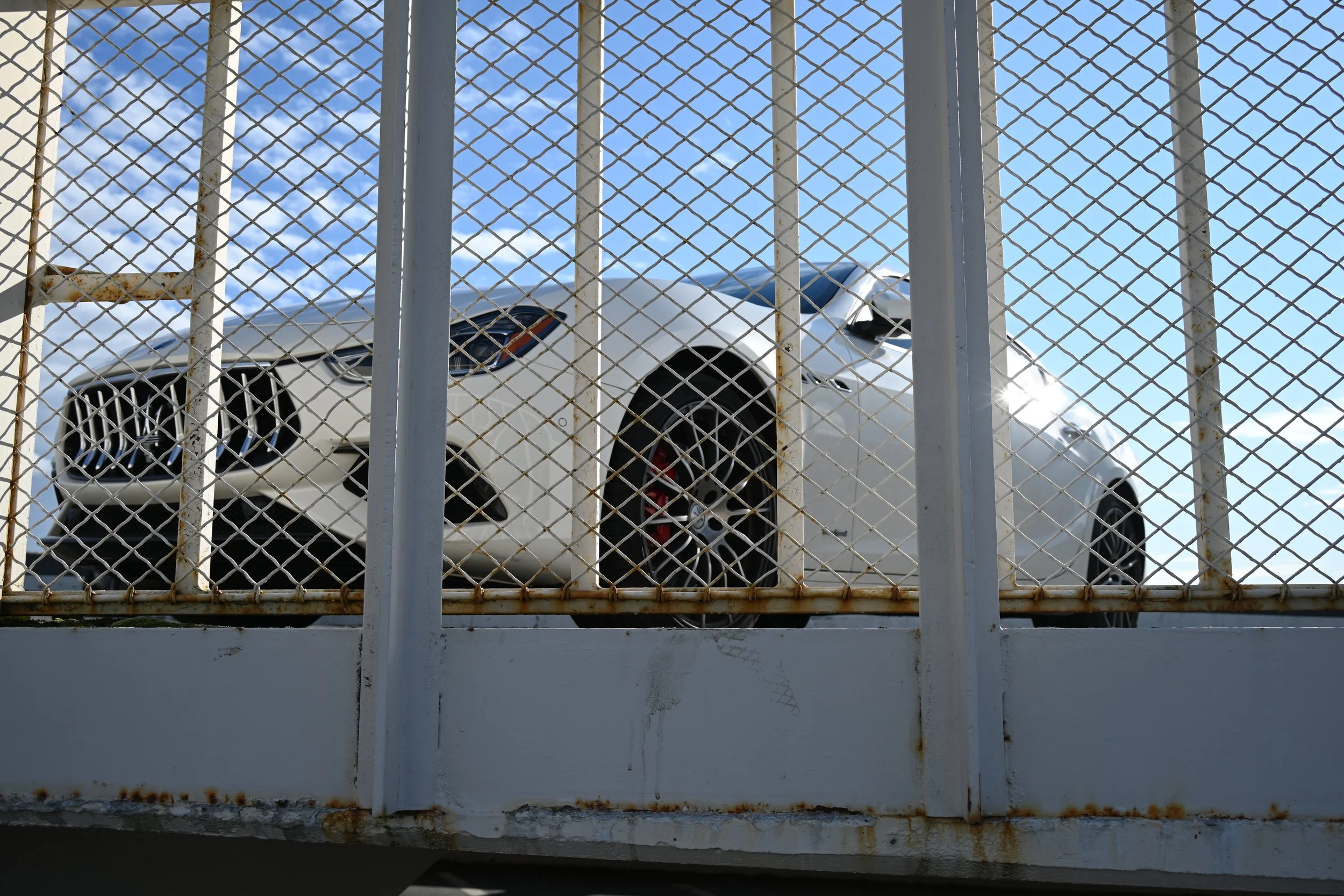 A white luxury sports car seen through a rusty metal fence on a bridge, with a blue sky and some clouds in the background.