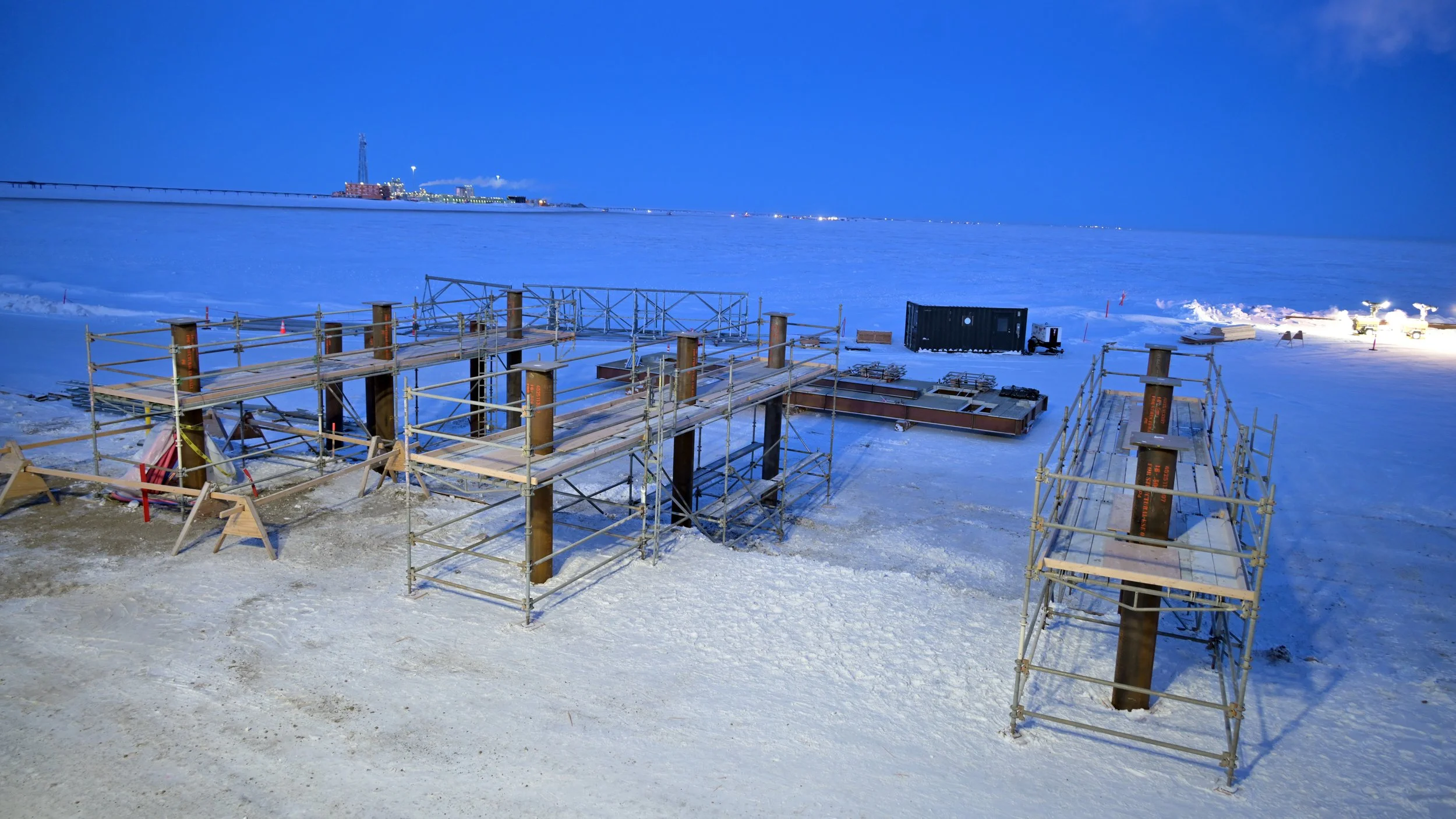 Construction scaffolding set up on snow-covered ground at dusk with a frozen body of water and industrial buildings in the background.
