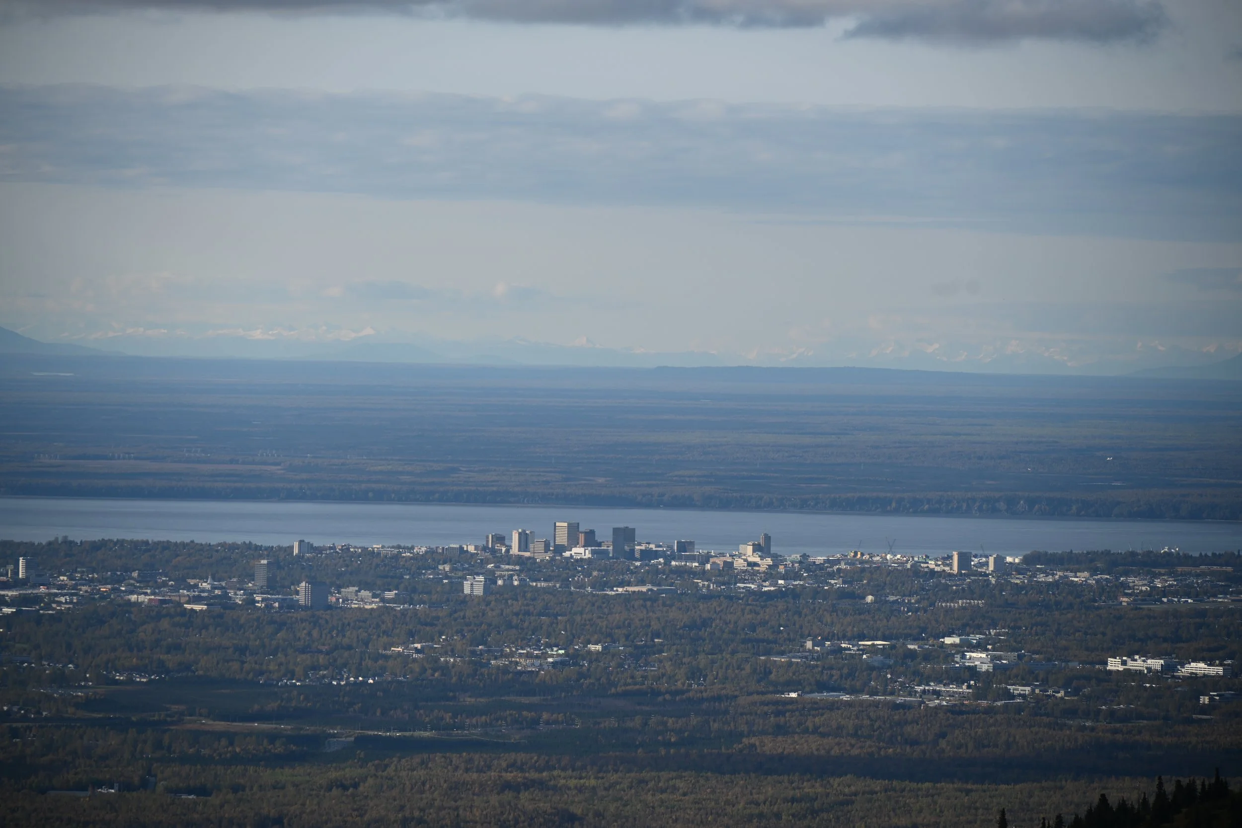 A panoramic view of a city near a large body of water with mountains in the background, under cloudy skies.