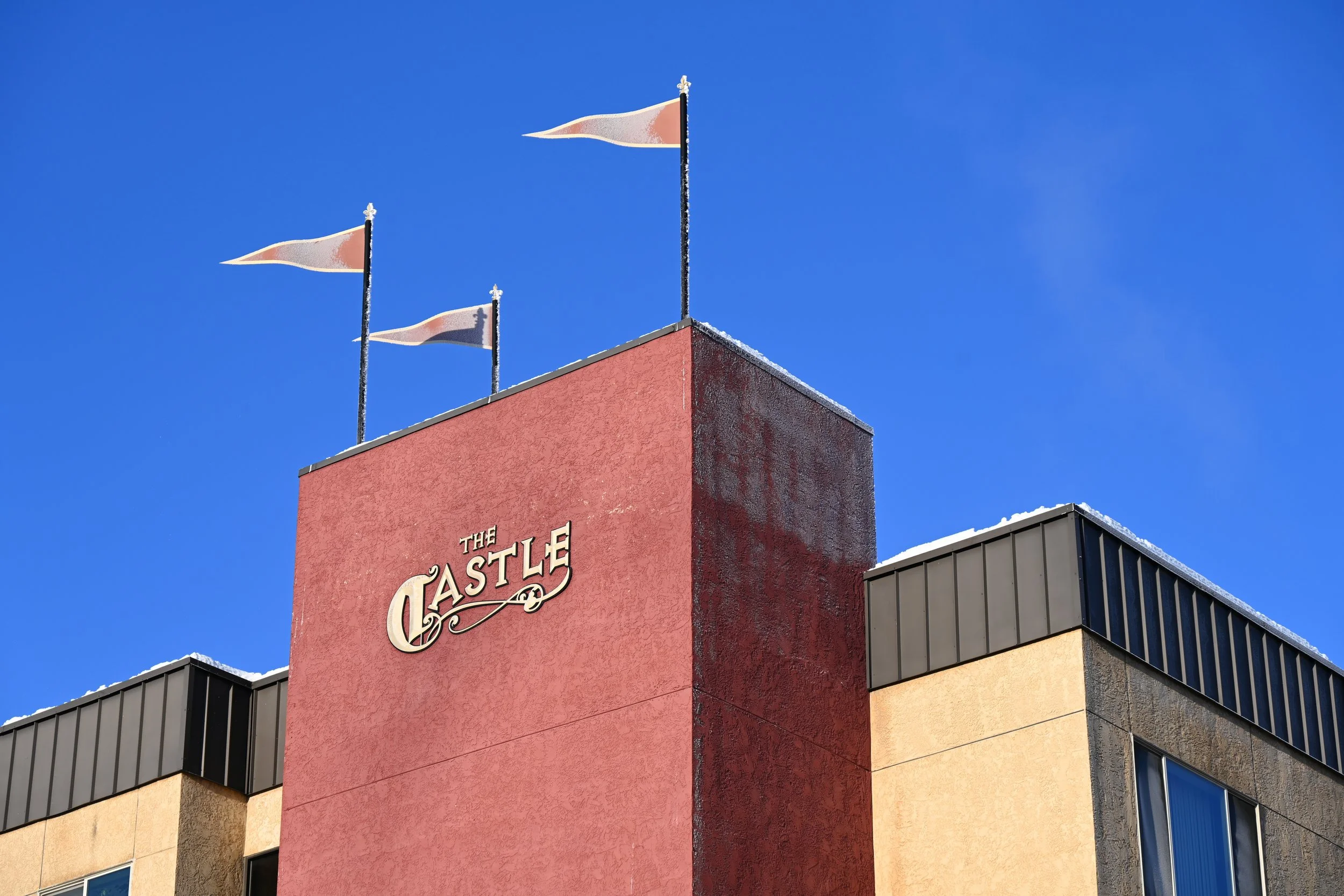 The upper corner of a building named 'The Castle' with three flags flying against a clear blue sky.