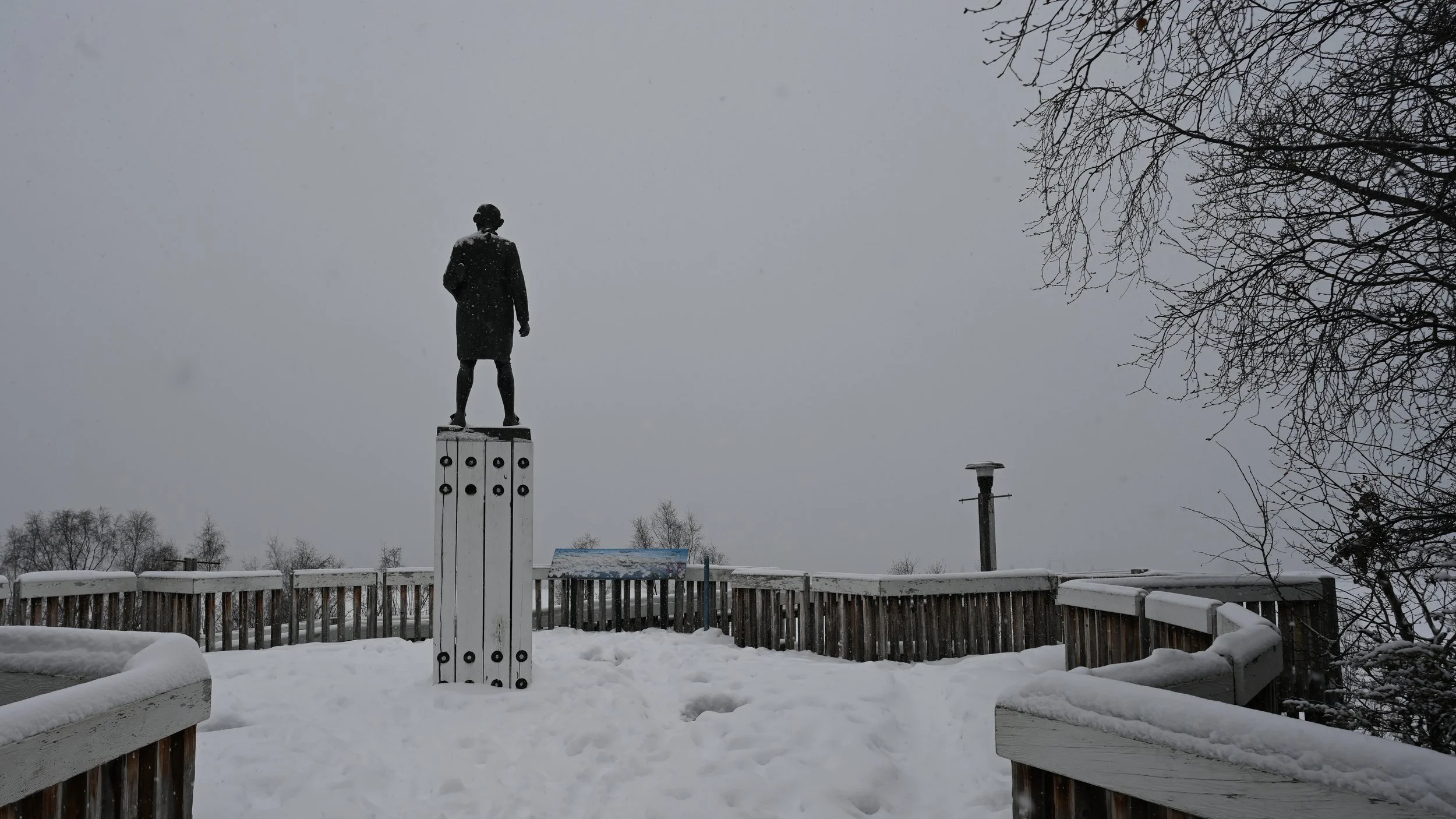 A statue of a person standing on a pedestal on a snow-covered deck, with a gray sky and bare trees in the background.