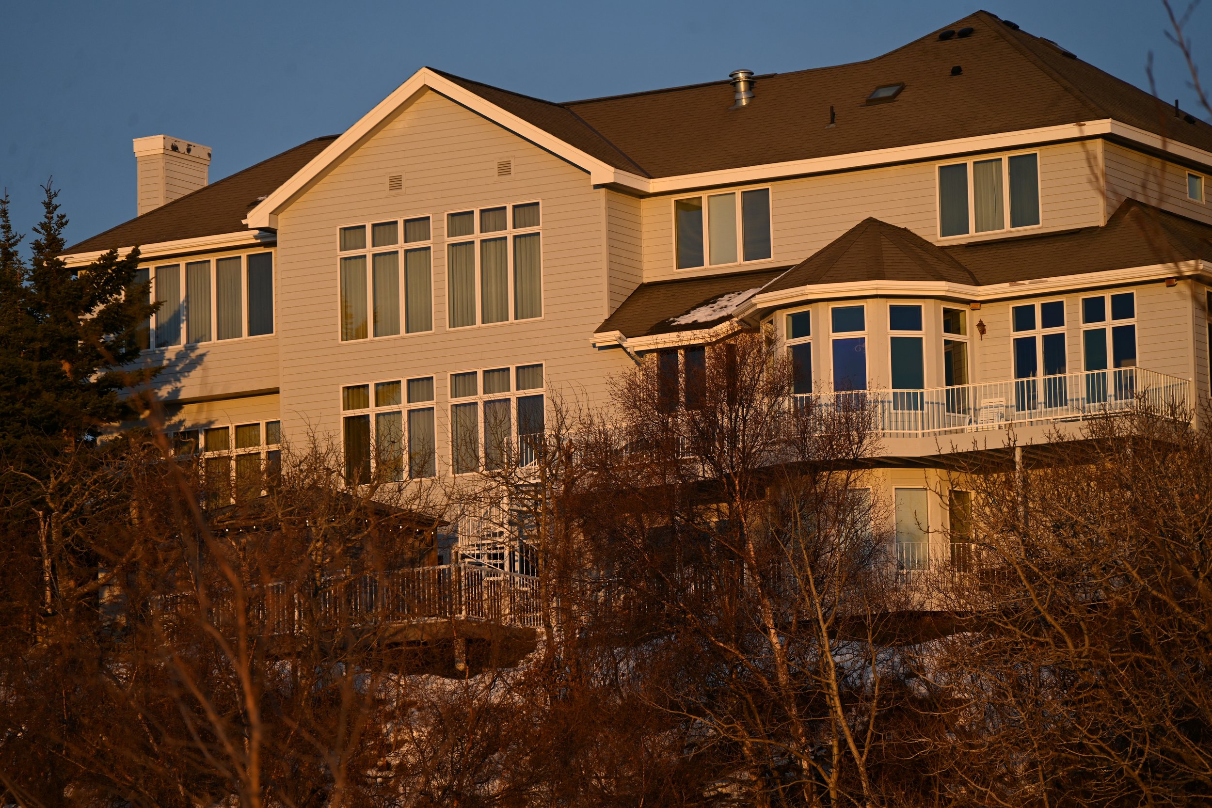 Large white multi-story house with multiple windows and balconies, surrounded by leafless trees, illuminated by warm sunset light.