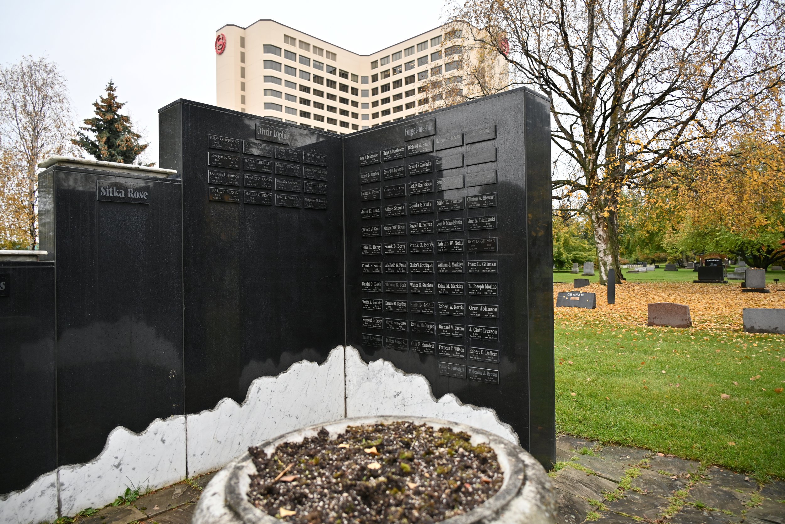 Memorial wall with engraved plaques outside in a cemetery, surrounded by trees with fall foliage and a high office building in the background
