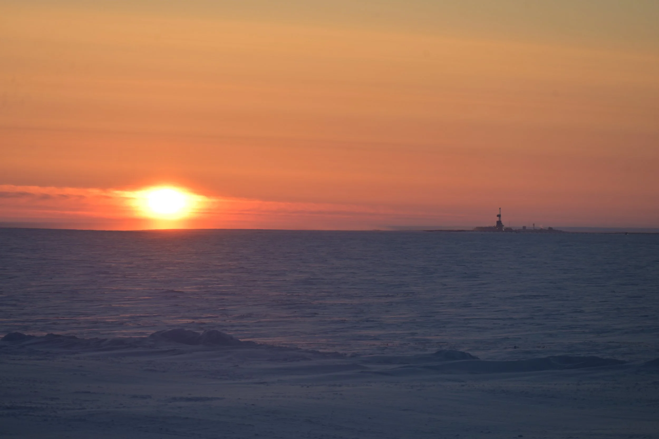 Sunset over the ocean with icy shoreline and a lighthouse in the distance.