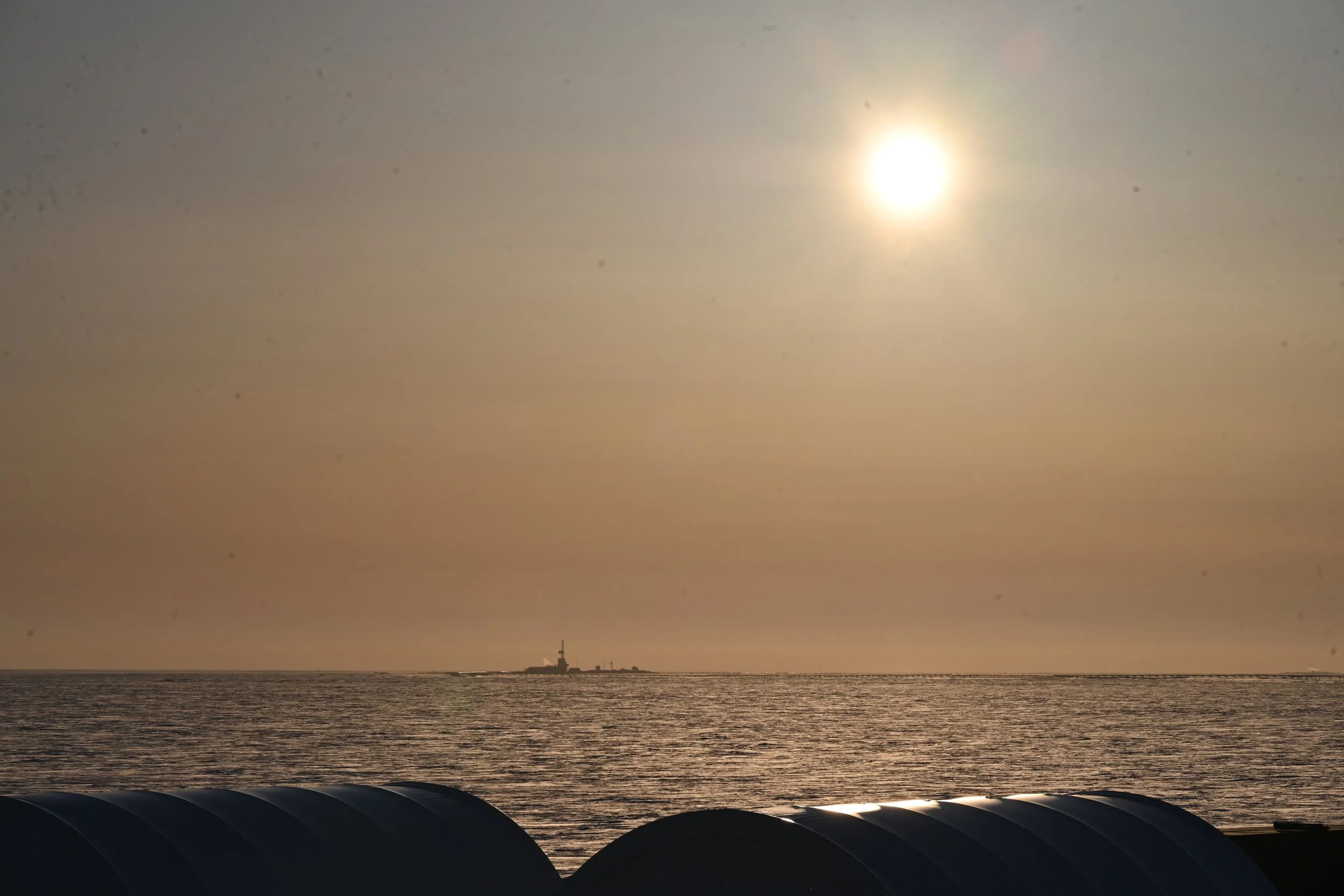 Sunset over the ocean with a lighthouse in the distance and large black pipes in the foreground.