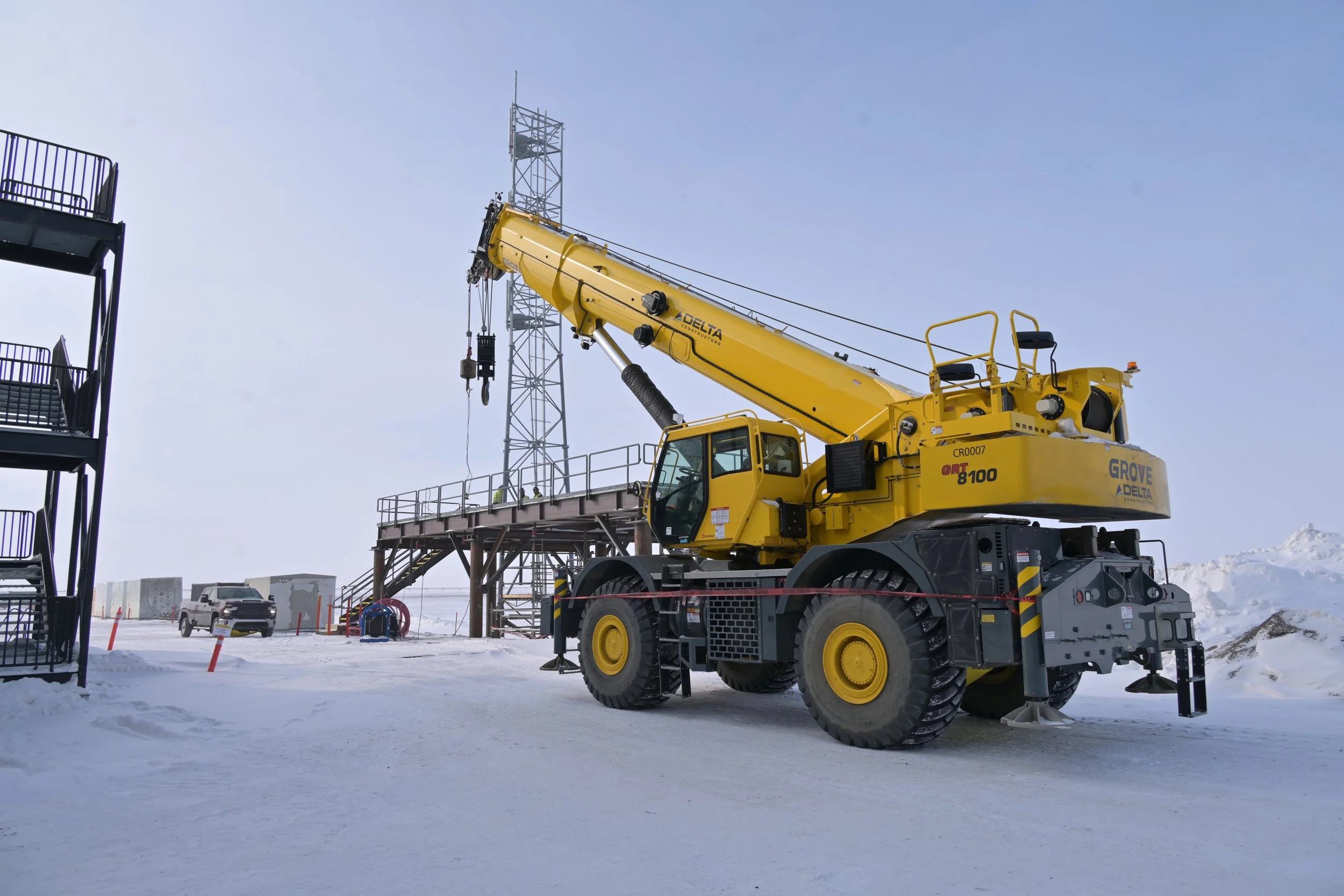 Yellow crane on snowy ground next to a metal platform and a tower, with snow-covered landscape in the background.