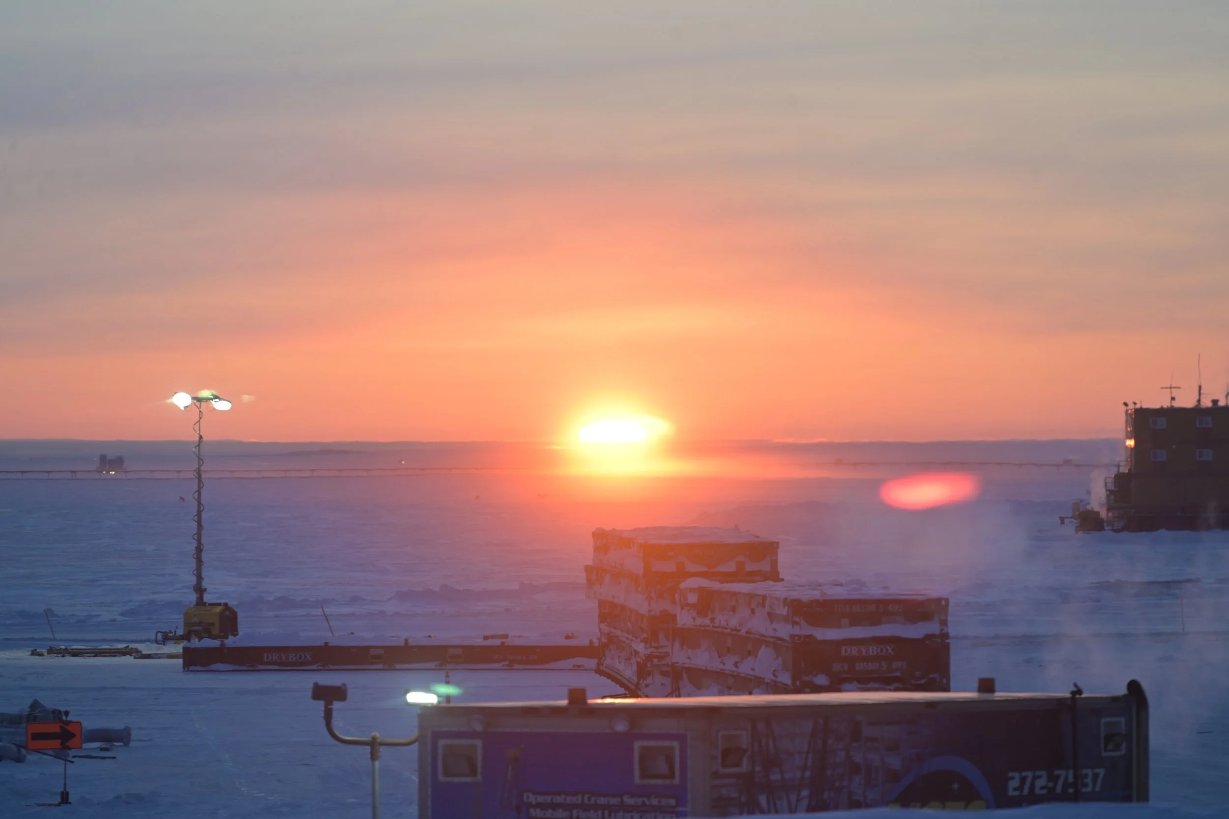 Sunset over a snowy landscape with industrial equipment and buildings, some illuminated by artificial lights.