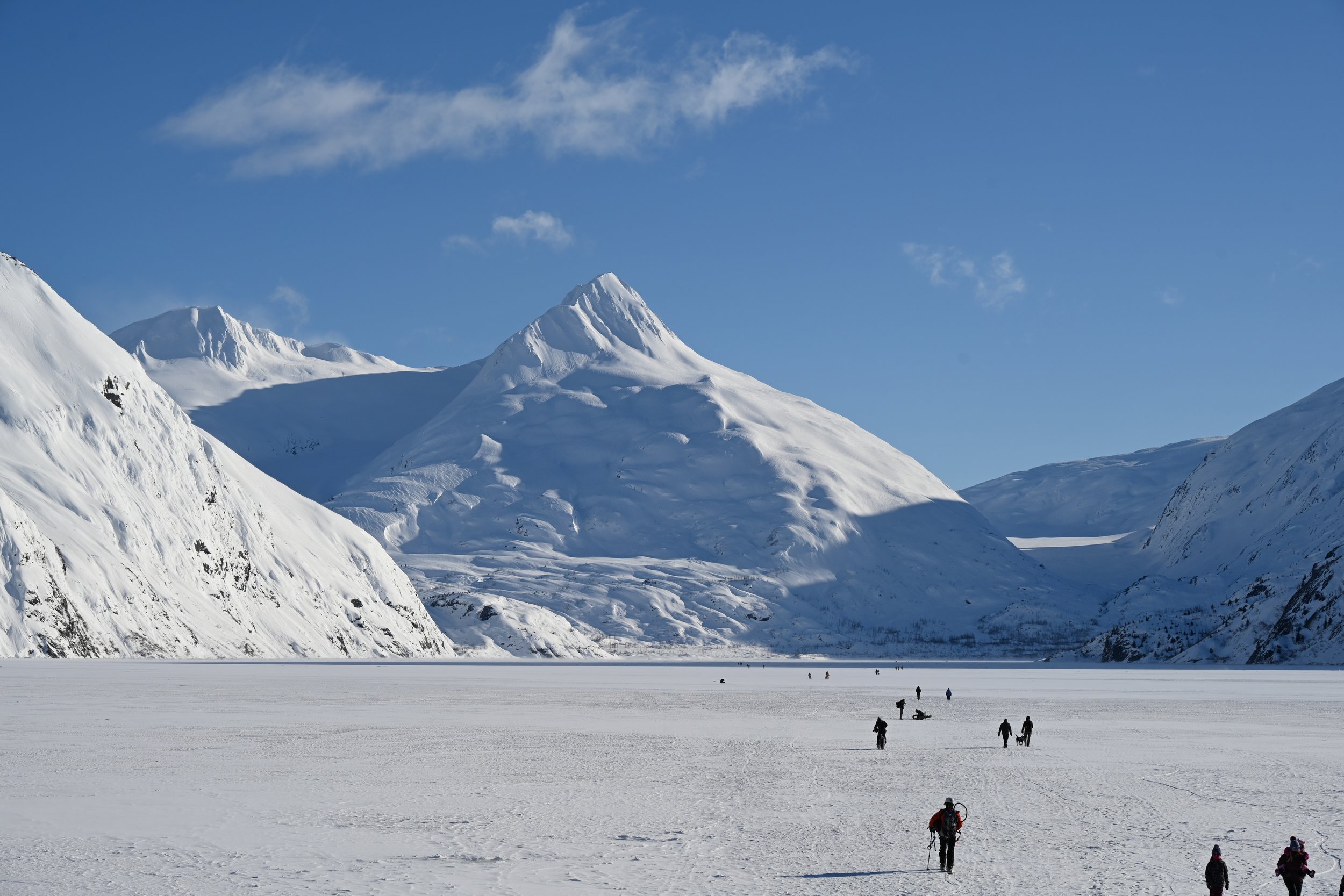 Snow-covered mountains with partly cloudy blue sky and people walking on a frozen landscape.