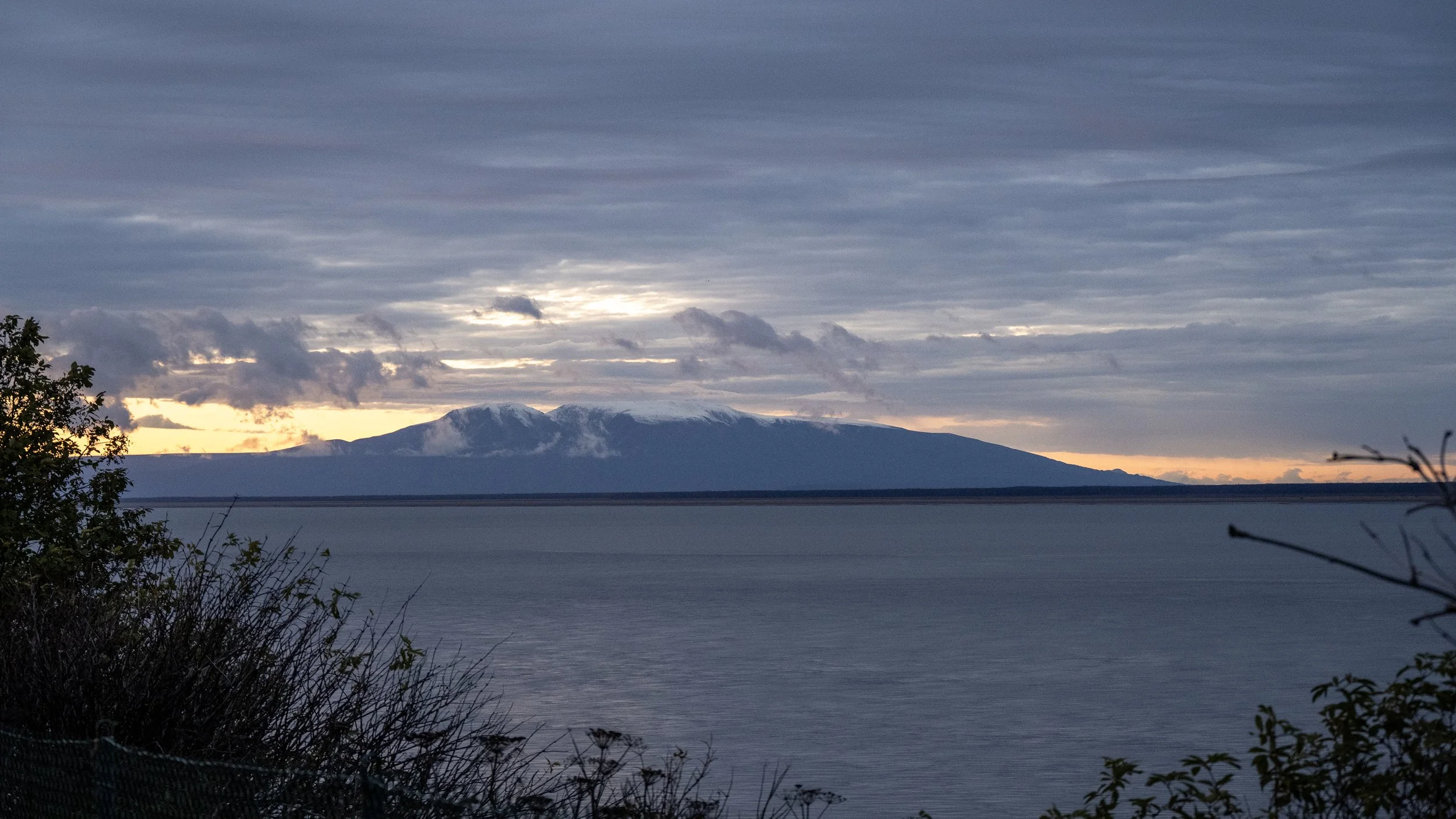 Scenic view of a mountain with snow-capped peaks in the distance, partially obscured by clouds, with a large body of water in the foreground and some bushes on the shore.