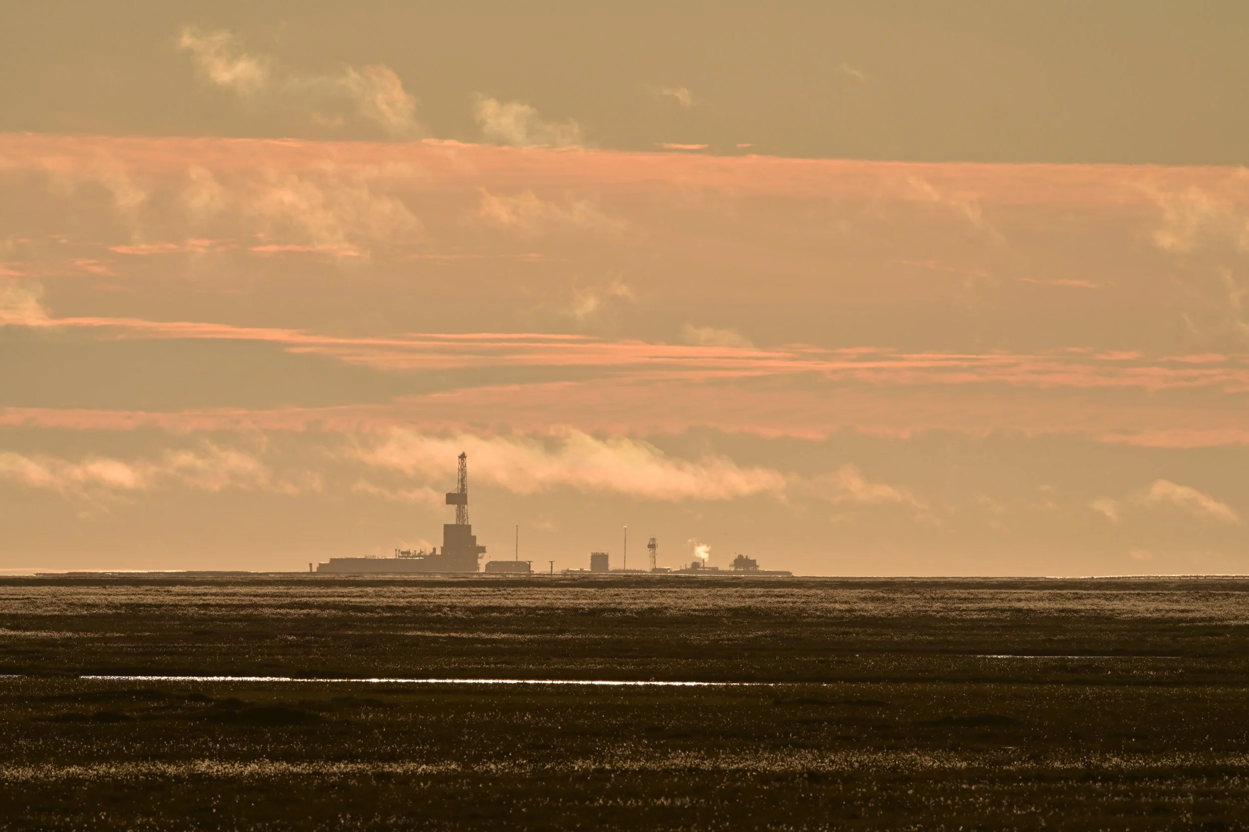 View of an offshore oil platform silhouetted against an orange and pink sky, with reflective water and land in the foreground.