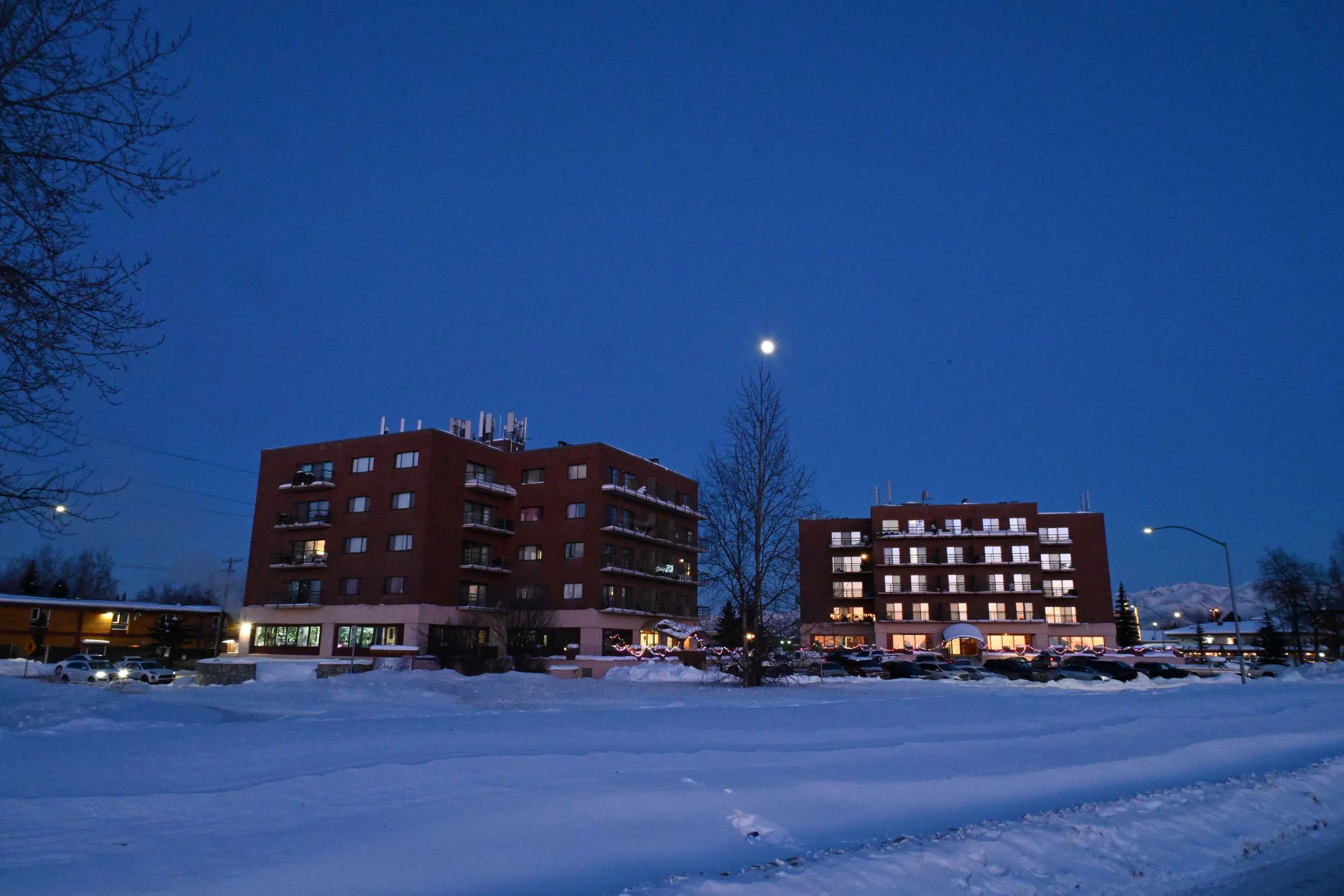 Nighttime view of two red-brick apartment buildings with lit windows, surrounded by snow with a clear blue sky, a bright moon, and a few visible stars.