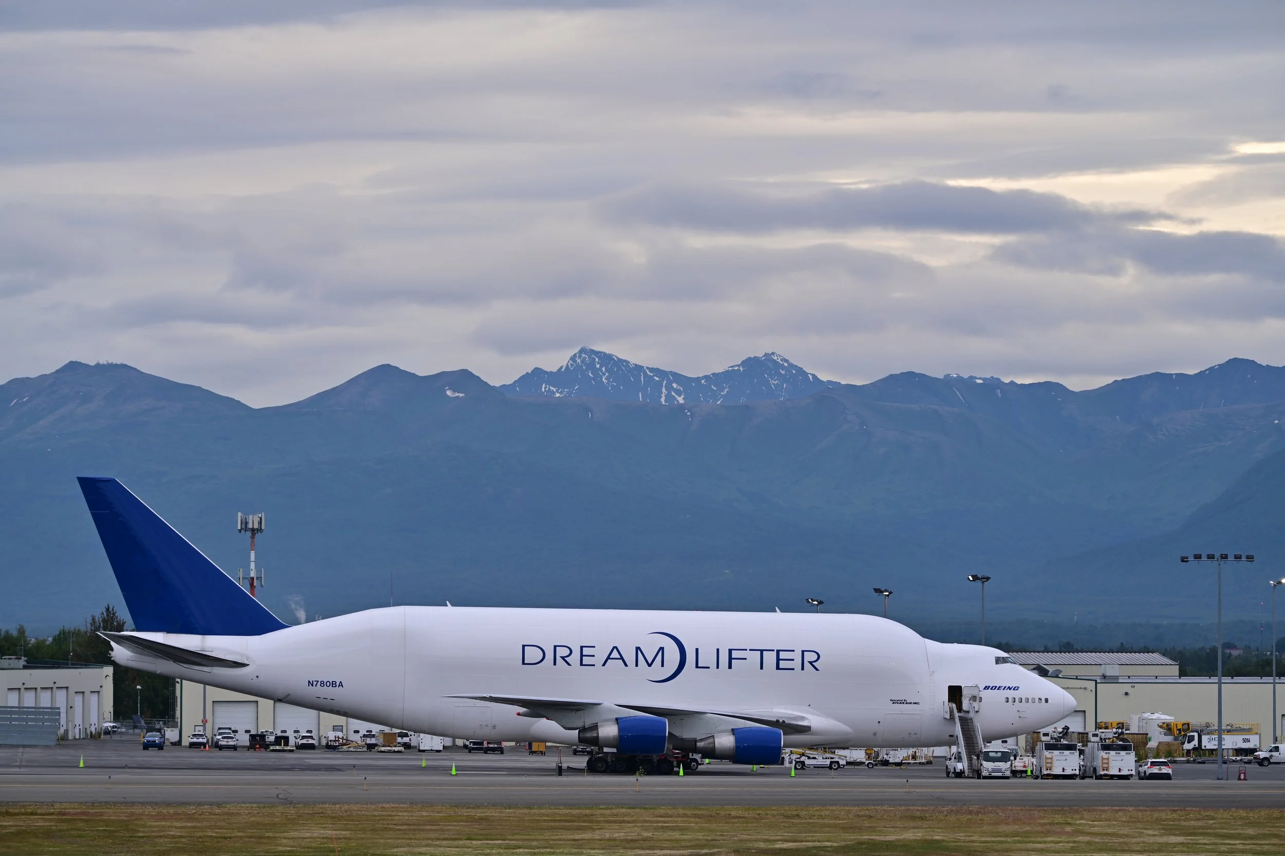 A Boeing DreamLifter cargo aircraft on the tarmac at an airport with mountains and cloudy sky in the background.