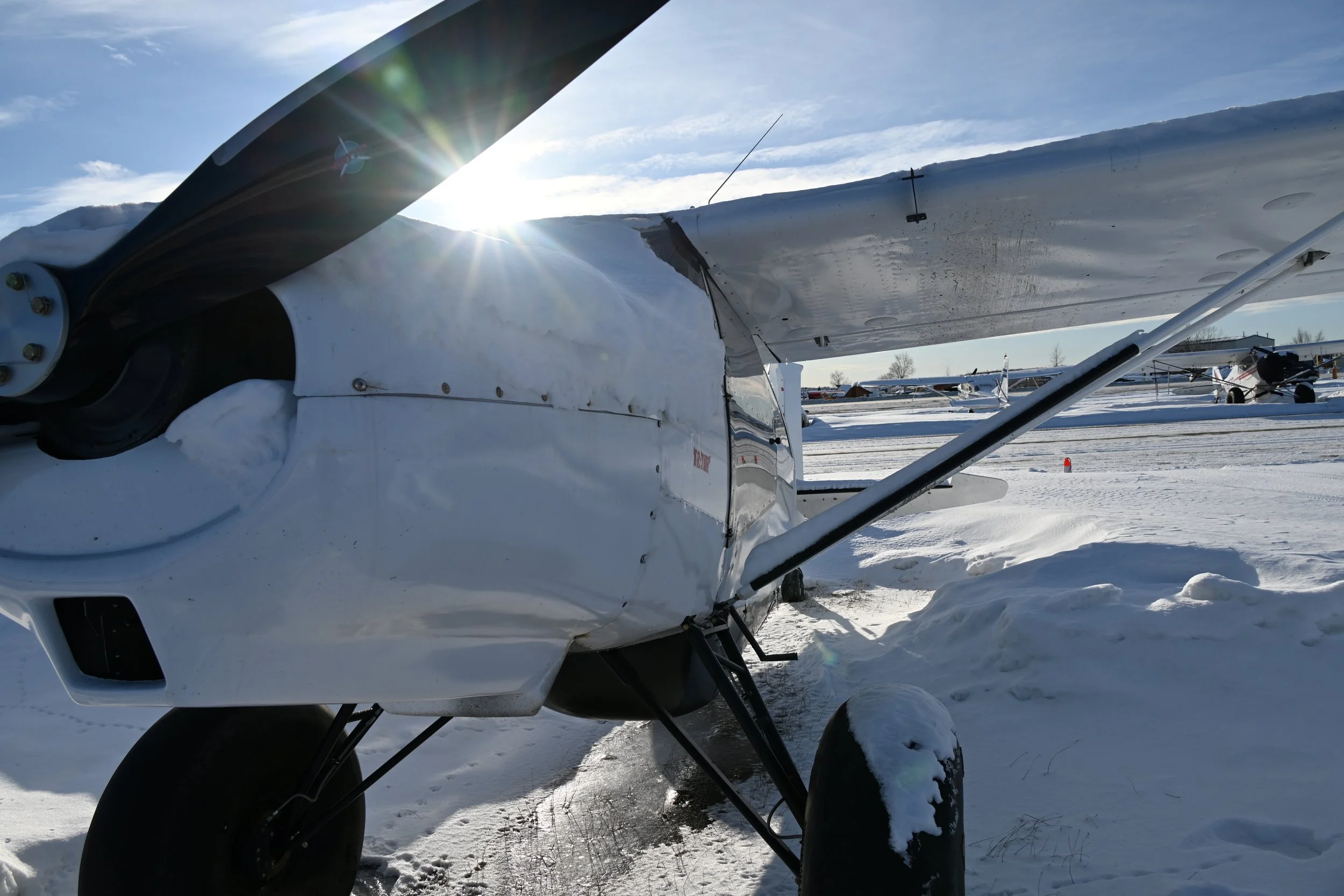 Small white airplane parked on snow-covered ground with other aircraft in the background, under a clear blue sky with the sun shining.
