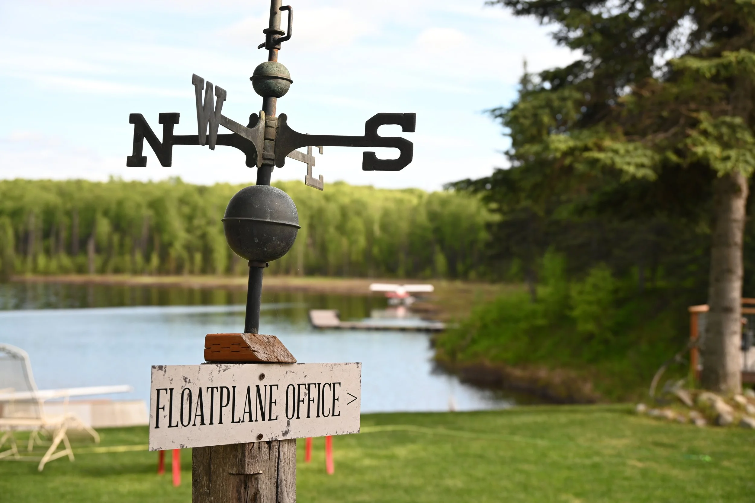 A weather vane with directions N, W, S, E mounted on a wooden post near a lake, with a white sign below reading 'Floatplane Office >'. In the background, there are trees, a grassy area, a dock with a floatplane, and a boat on the water.