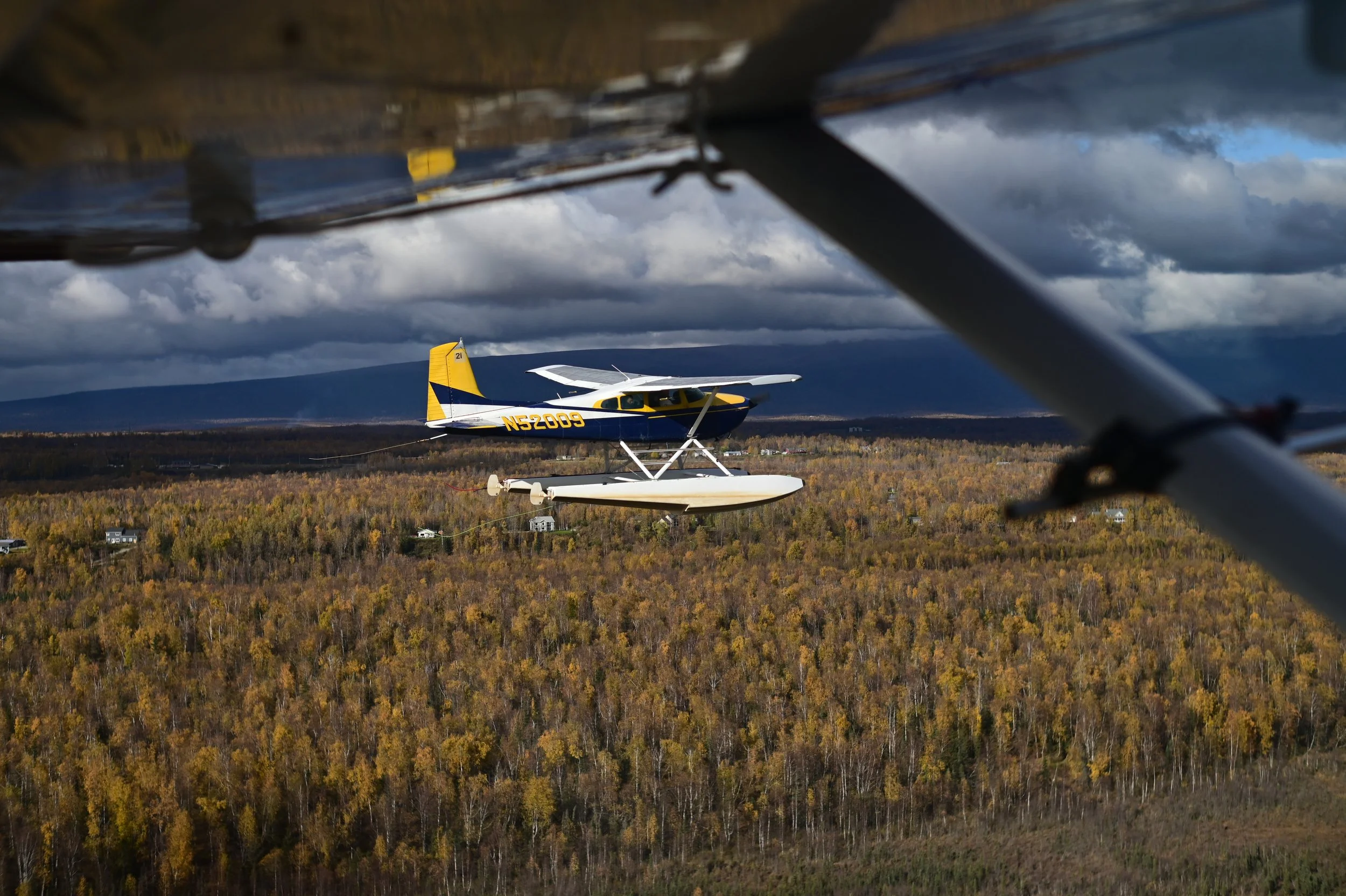 A seaplane flying over a landscape of trees with dark clouds overhead.