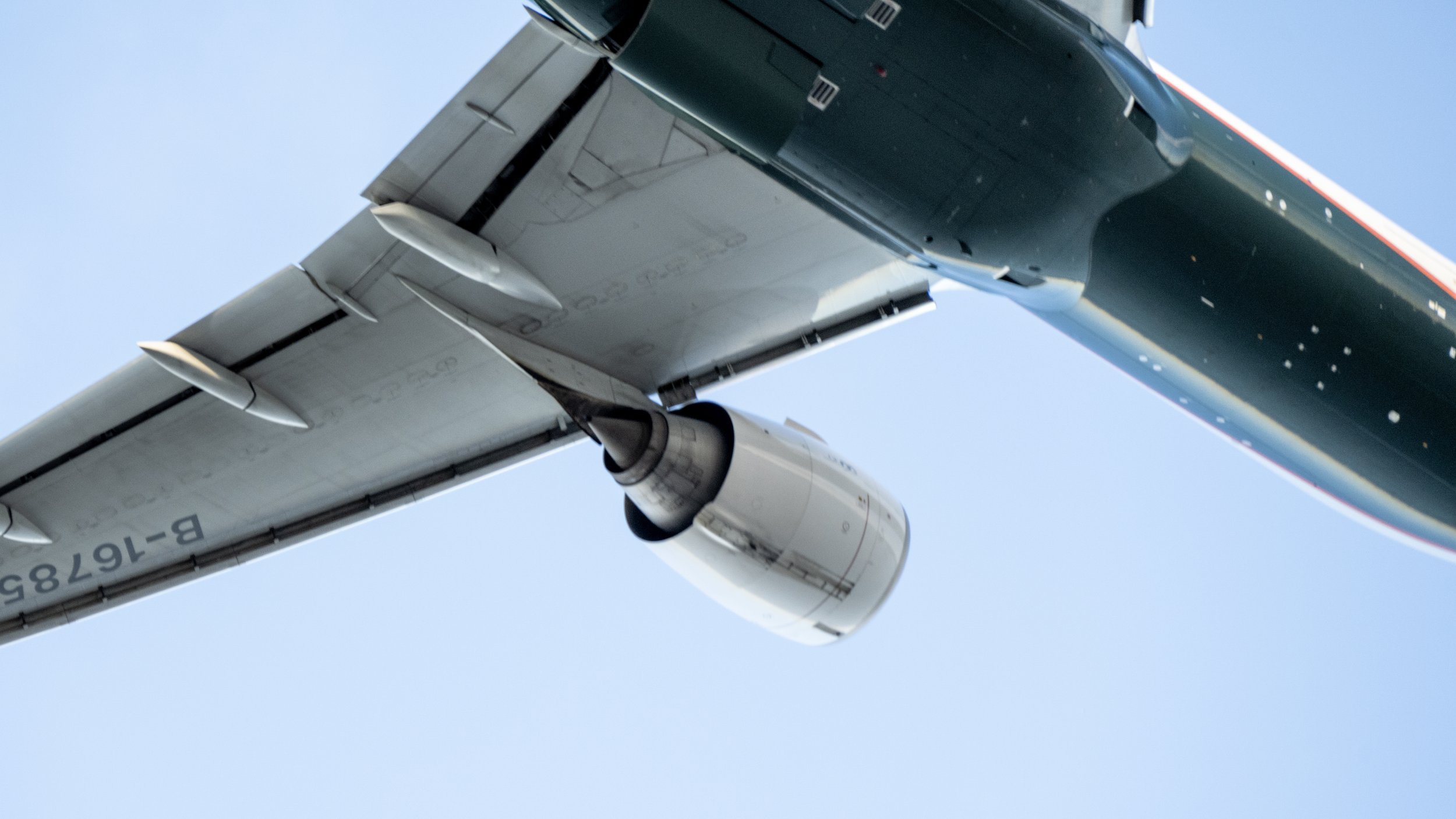 Close-up of an airplane's underside showing a white jet engine attached under the wing against a clear blue sky.