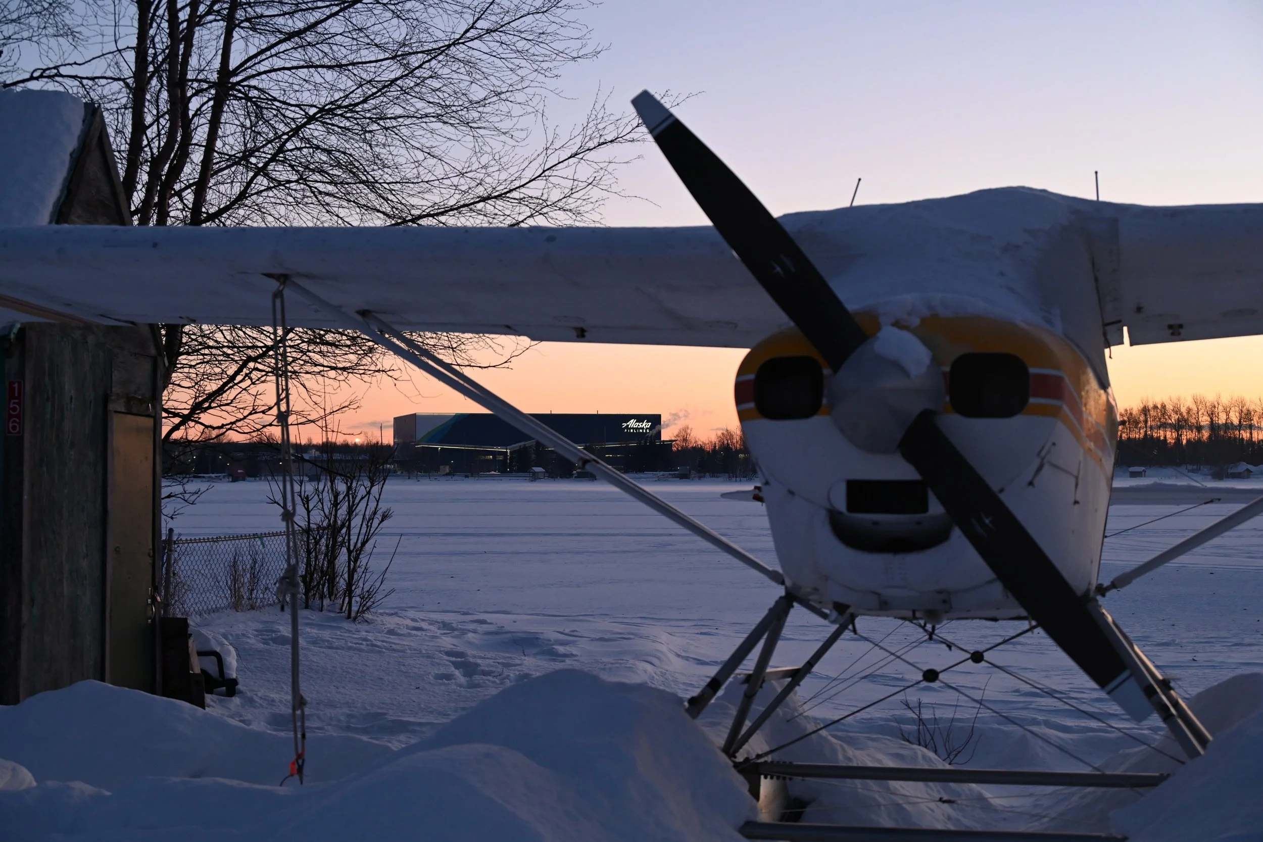 Small airplane parked on snow-covered ground with sunset sky in the background, trees, and a building labeled 'Alaska Airlines'.