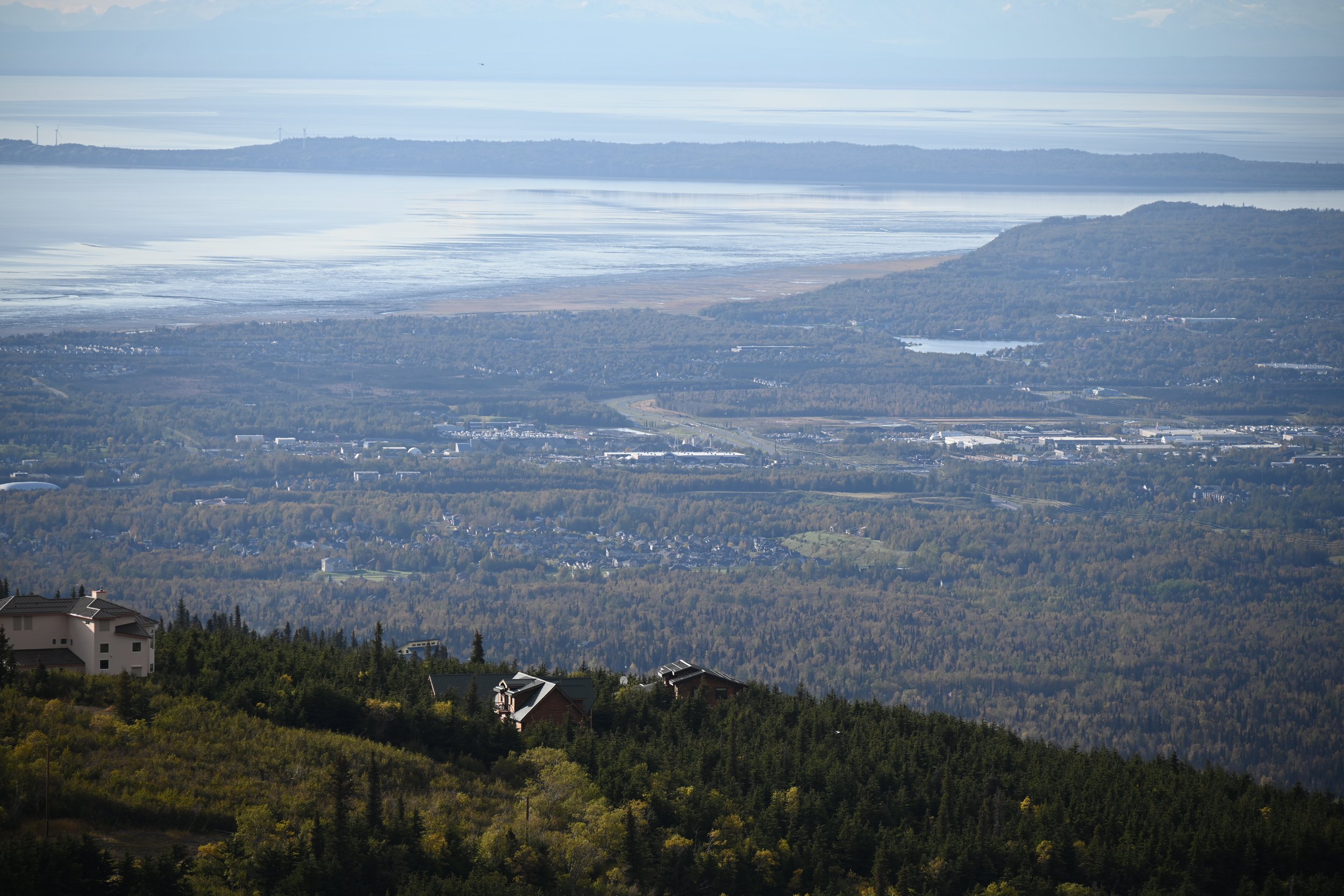 A scenic view of a hilly landscape with houses and dense forests in the foreground, a river and flatlands in the middle ground, and a large body of water with an island in the background under a partly cloudy sky.