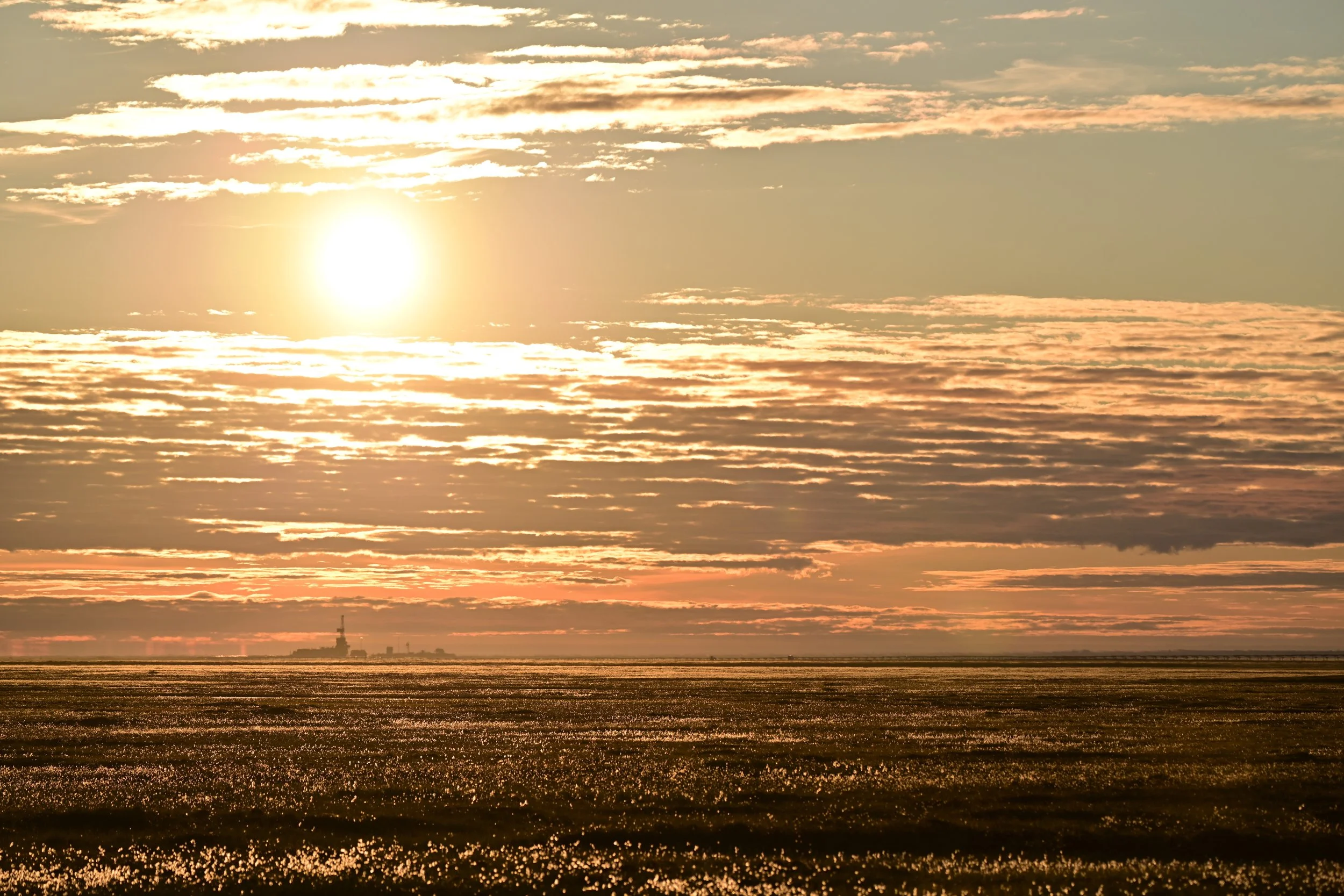 Sunset over a flat field with a distant offshore oil rig or platform on the horizon, partially covered with clouds.