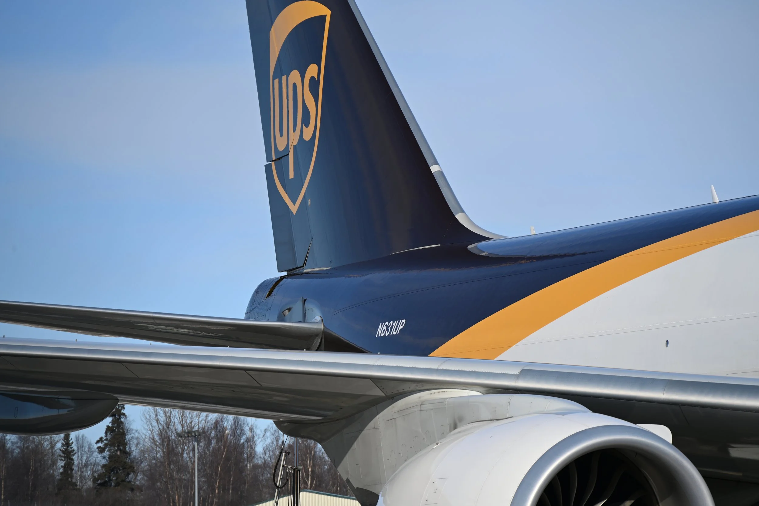 Close-up of the rear section of a UPS delivery airplane, showing the UPS logo on the vertical stabilizer, part of the wing and jet engine, with trees and a clear blue sky in the background.