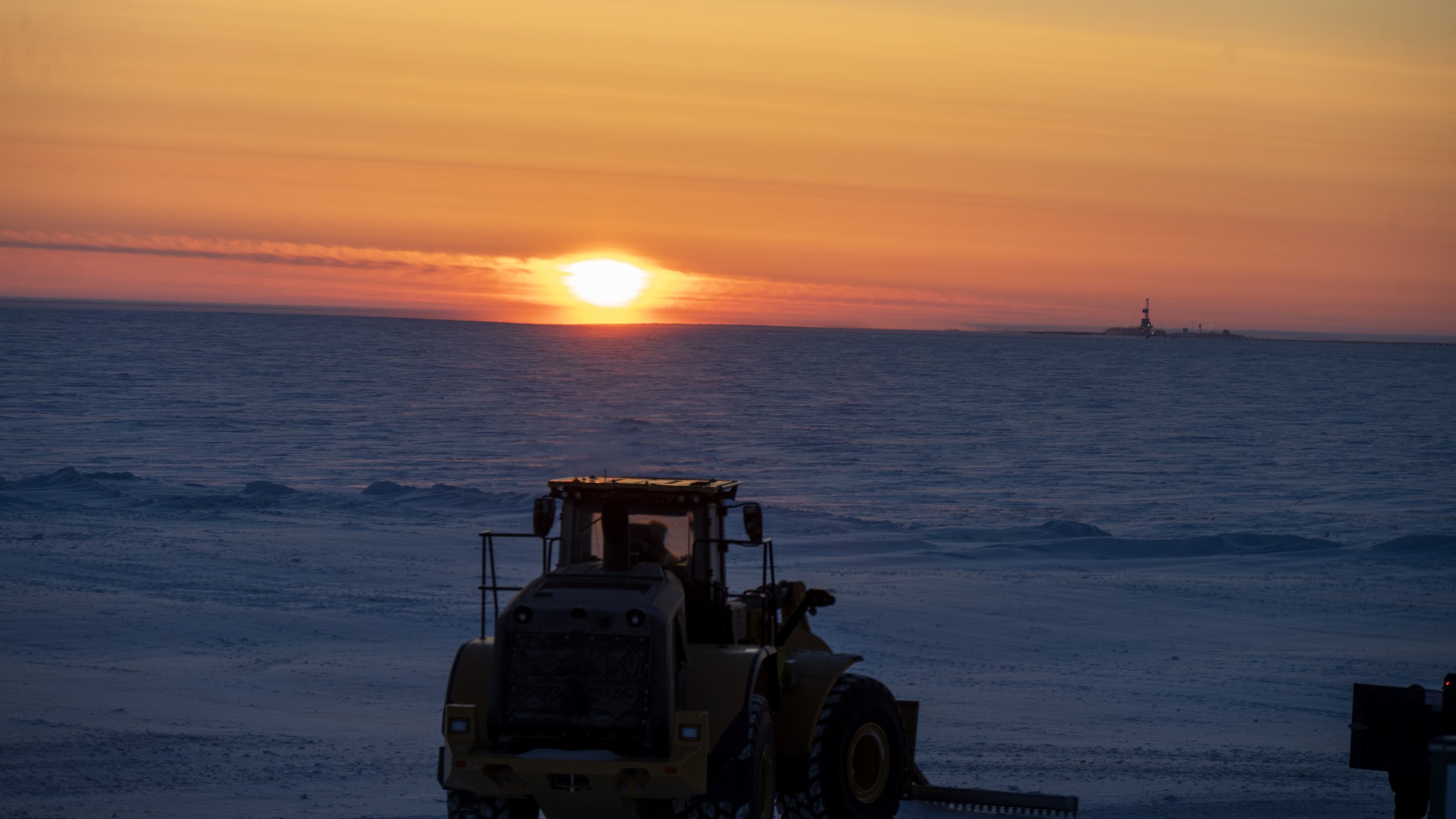 Sunset over icy ocean with a lighthouse on the horizon, a snowplow in the foreground, and a person nearby.