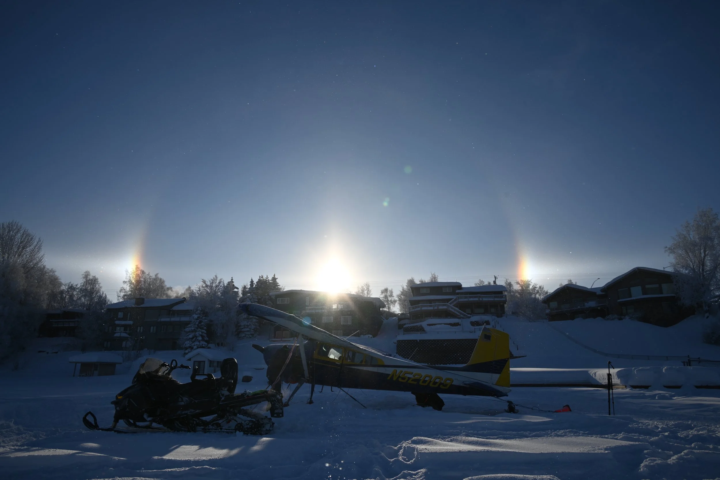 A snow-covered landscape at night with a small plane and snowmobile in the foreground, houses on a hillside in the background, and moonlight creating a halo and sundogs in the clear sky.
