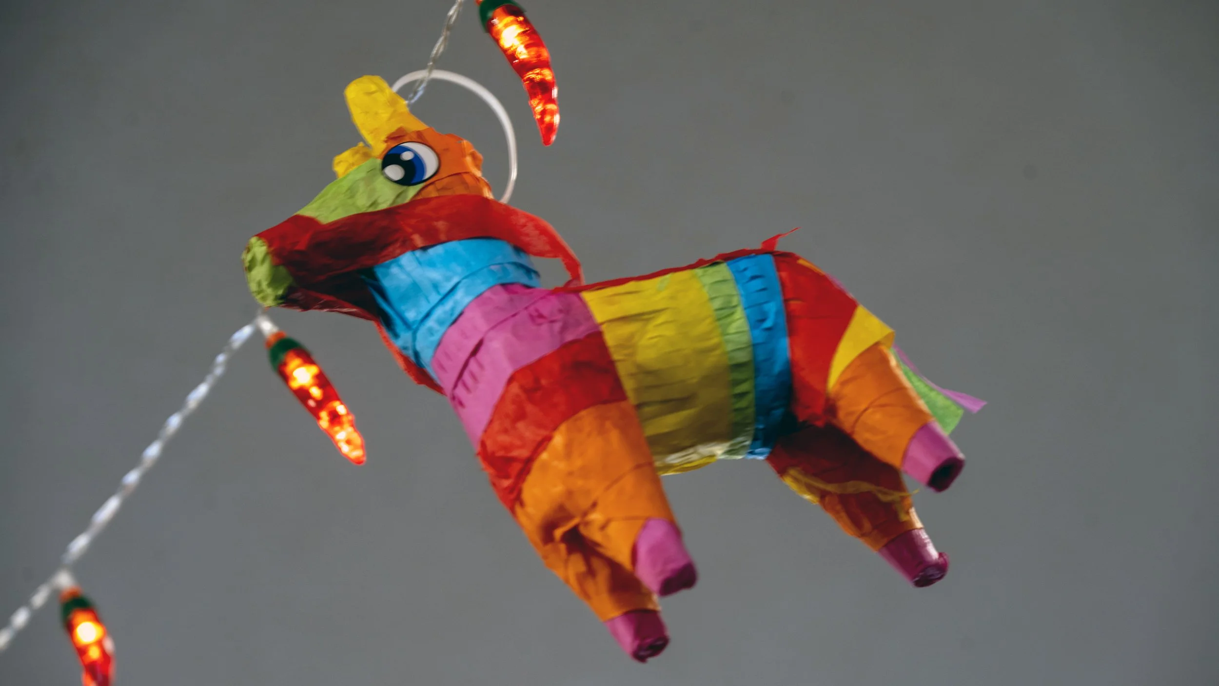 Colorful piñata in the shape of a unicorn, decorated with rainbow stripes and LED lights, hanging against a gray background.