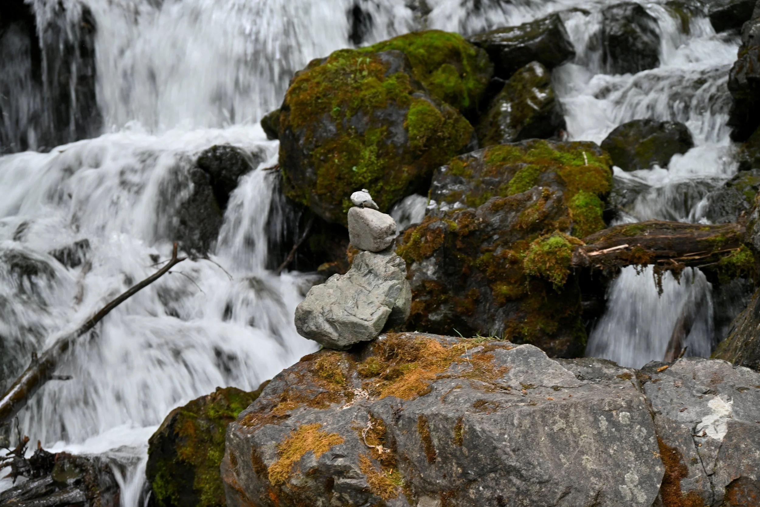 Stacked rocks on a moss-covered rock near a flowing creek in a natural setting.