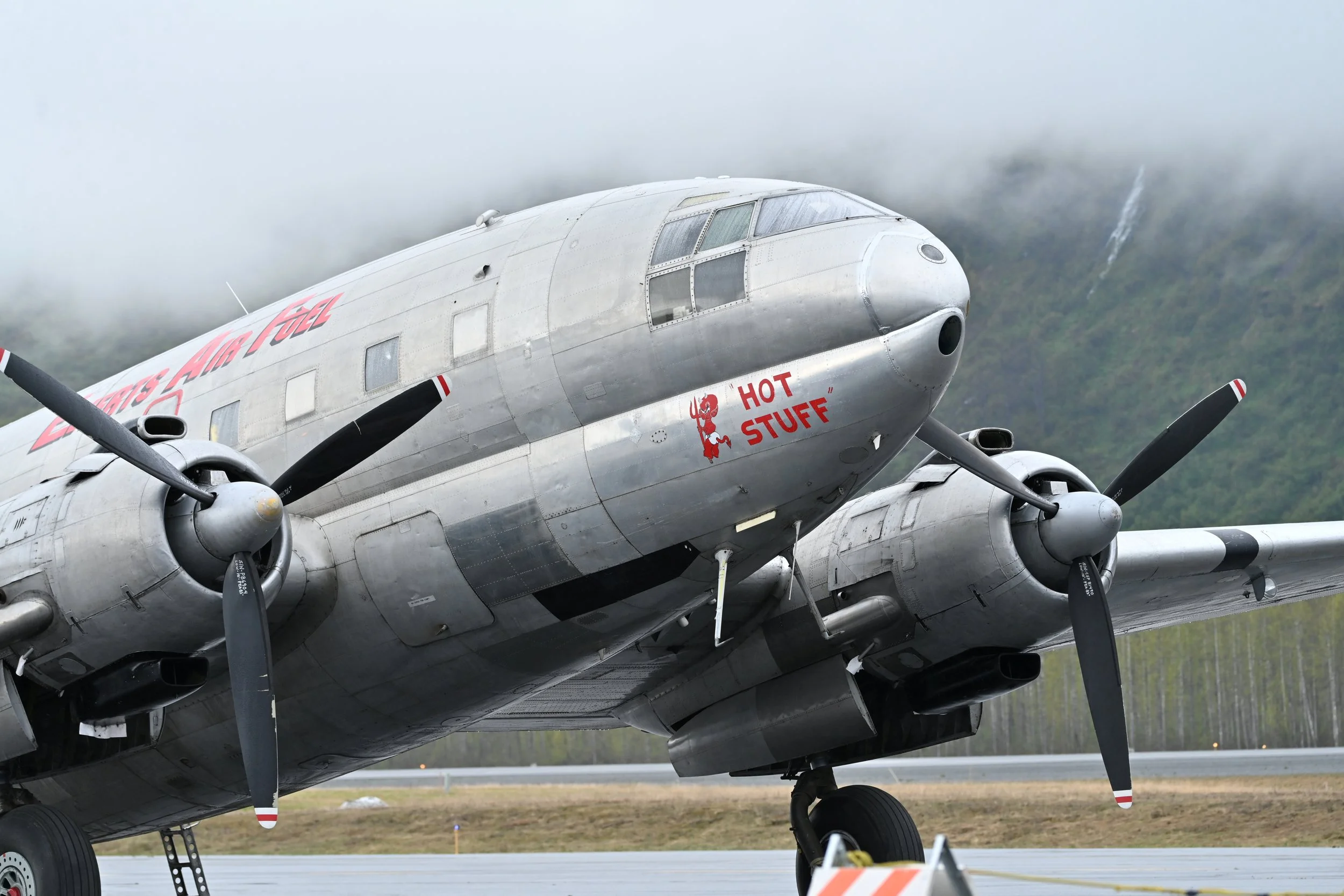 A vintage propeller airplane on the runway with a mountain and cloudy sky in the background. The nose of the plane has red military-style lettering and a cartoon devil with the words "HOT STUFF".