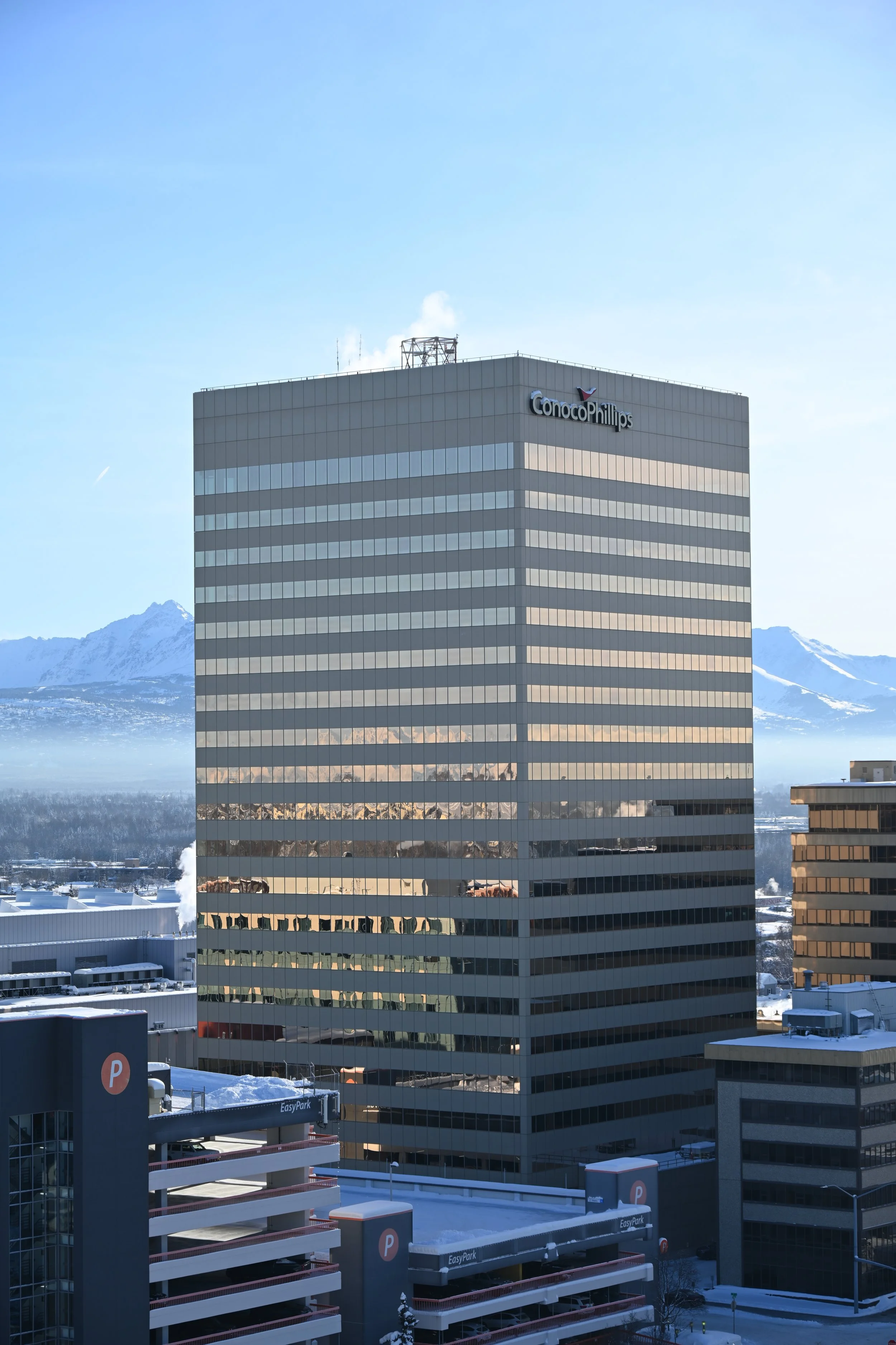 Tall office building with a ConocoPhillips logo, surrounded by other buildings and snow, with mountain range in the background.