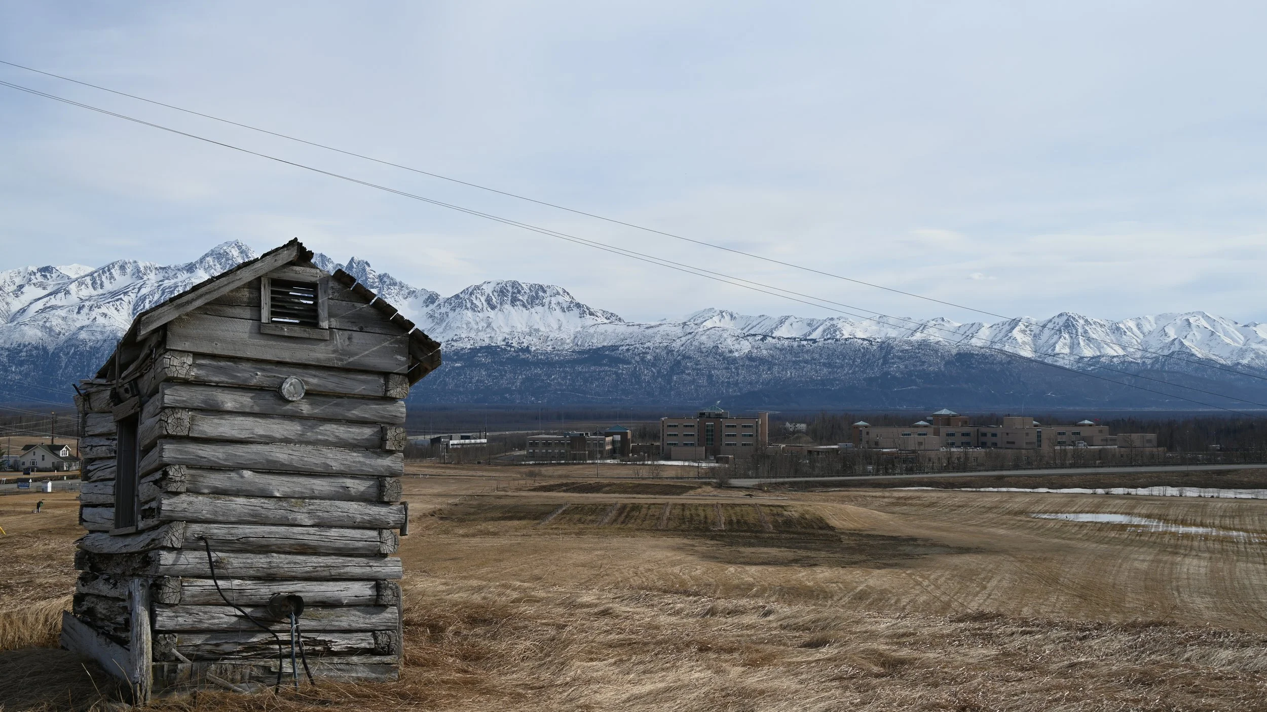 A weathered old wooden shed in a rural field with snow-capped mountains in the background and a few modern buildings in the distance.