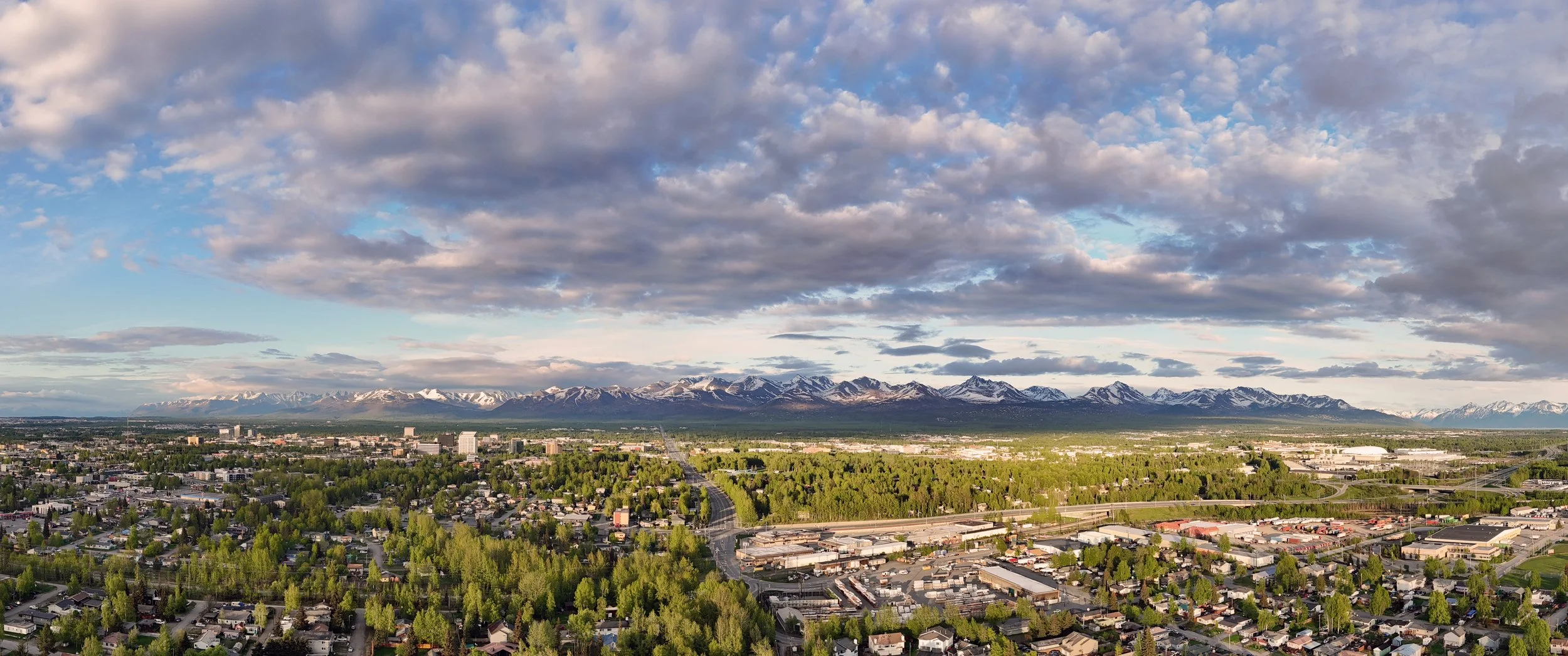 Aerial view of a city with residential and commercial areas, green trees, roads, and a mountain range with snow-capped peaks in the background under a partly cloudy sky.