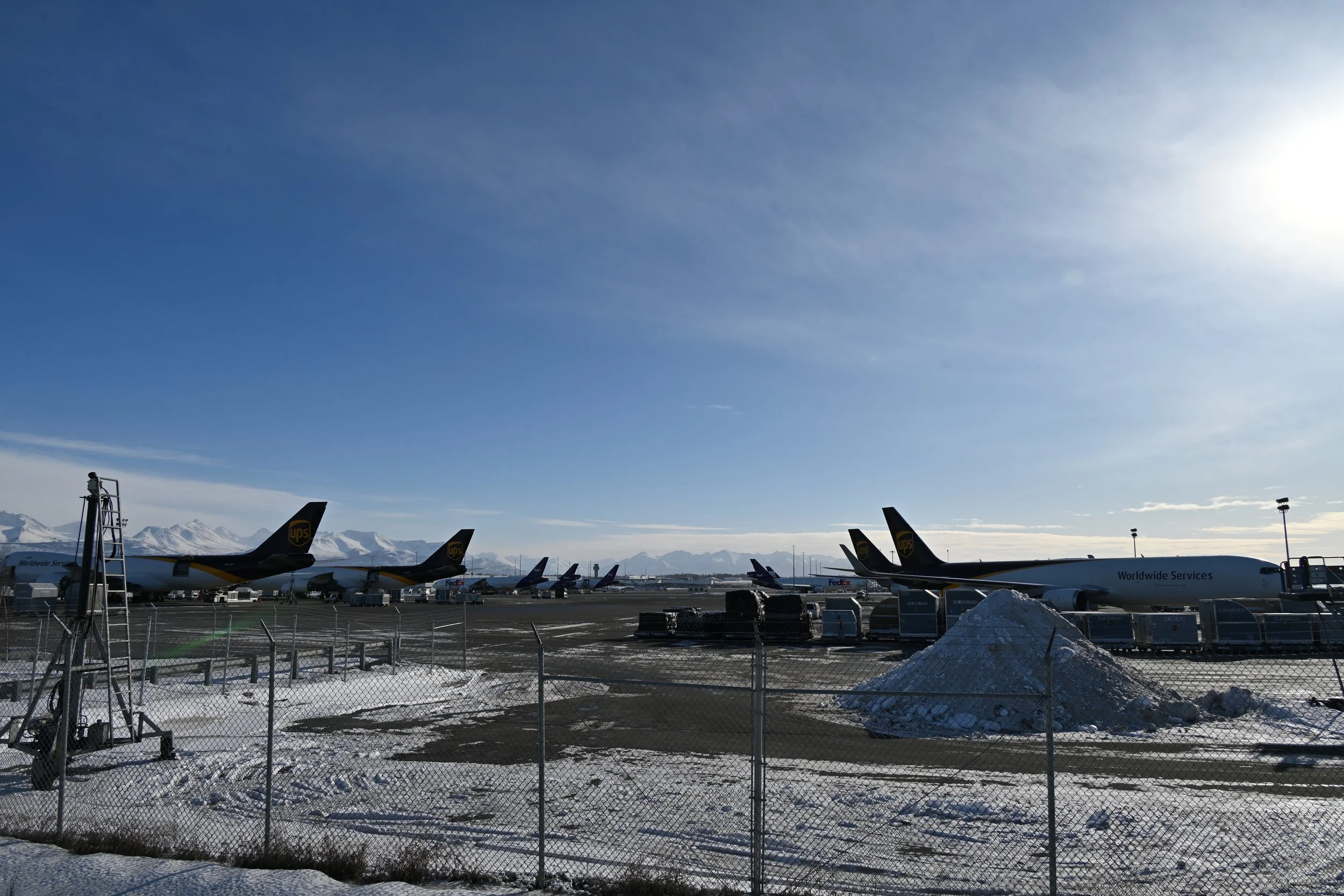 Aircrafts parked on snow-covered airport tarmac under a clear blue sky, with mountains in the background and snow piles in the foreground.