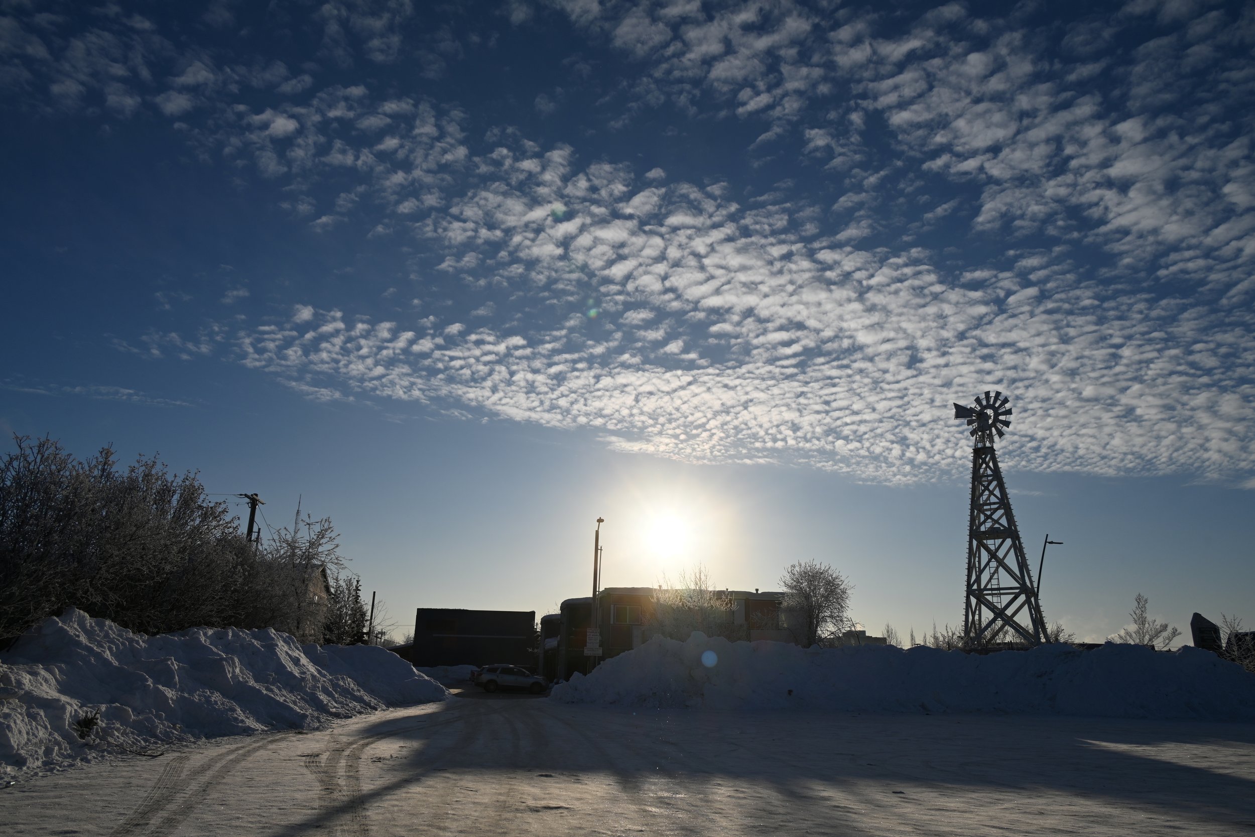 Snow-covered road leading to buildings and a windmill under a partly cloudy sky during sunset or sunrise.