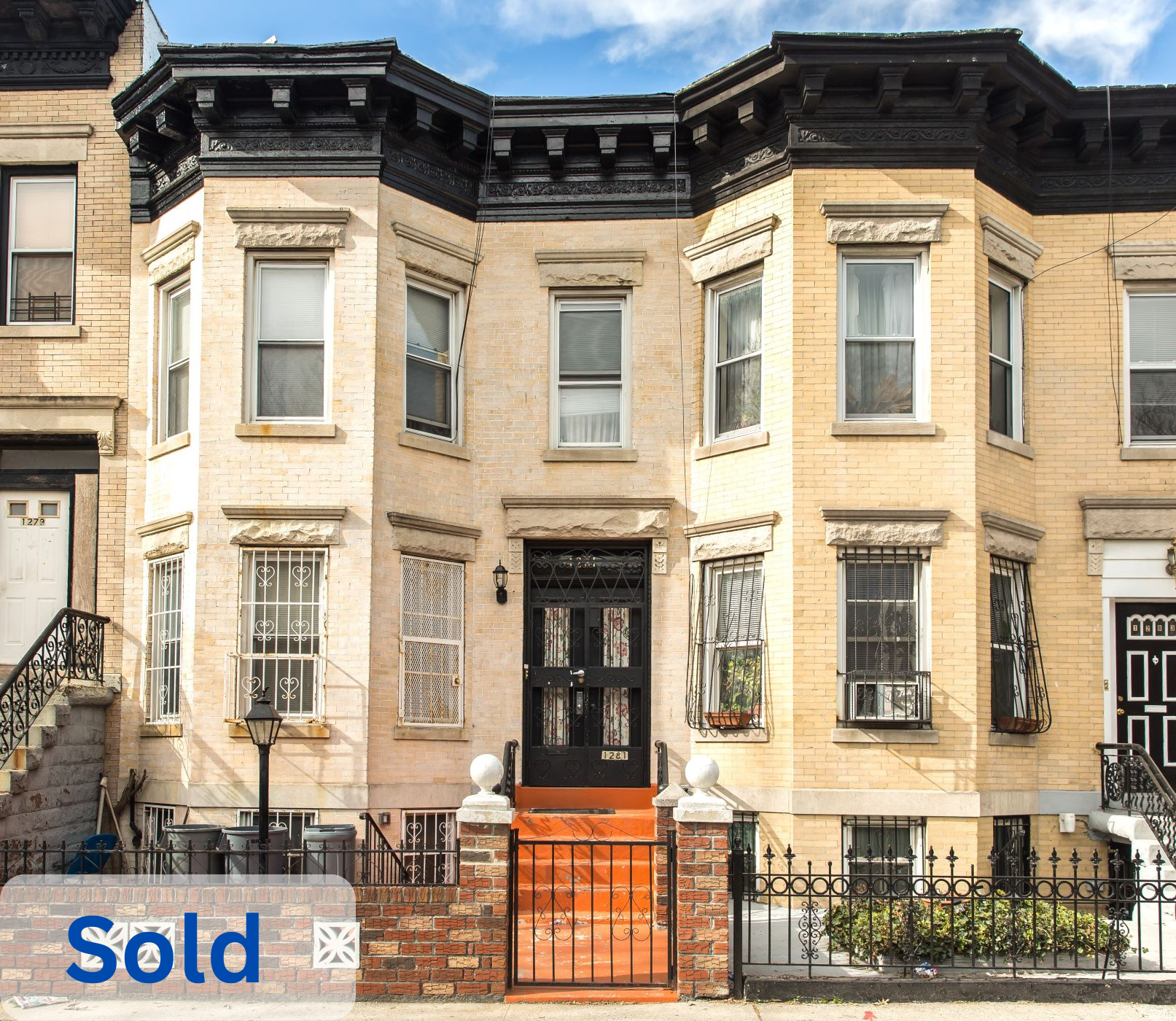 A three-story beige brick row house with black trim and an ornate black front door, located on a city street. The house has tall windows with decorative stone lintels, and small iron balconies on the second and third floors. The front yard has a brick and wrought iron fence, stairs with orange steps leading to the front door, and a small garden area. The word "sold" is overlaid in blue text at the bottom left corner.