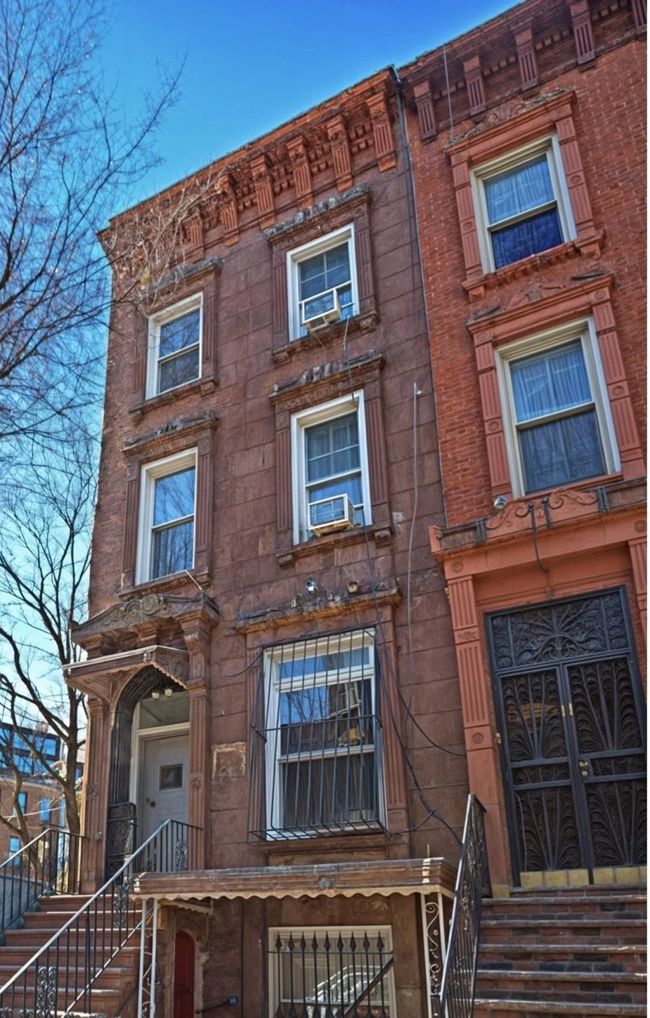 Brownstone apartment building with five windows, some with air conditioning units, decorative window frames, black iron stairs, and iron gate, with a tree and blue sky in the background.