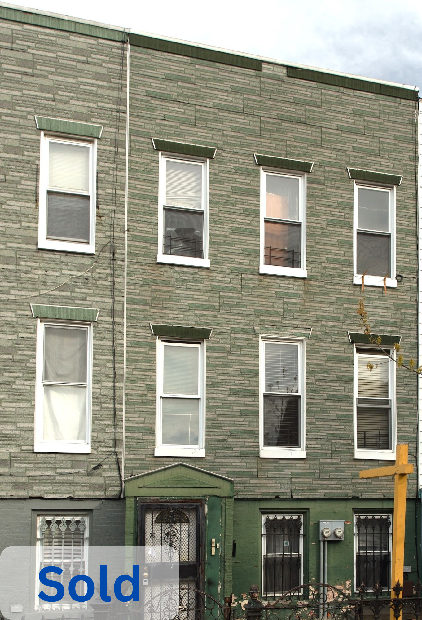 A four-story green brick building with eight windows, some with open or partially covered blinds, a black ornate front door with a security gate, and a small fenced front yard. The word "Sold" appears in blue in the lower left corner.