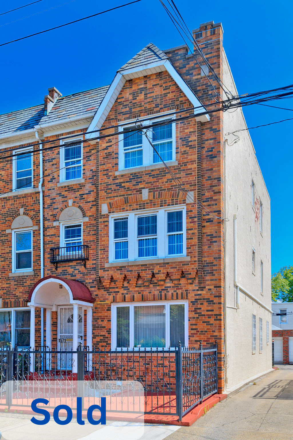 Brick apartment building with white window frames, small balcony, black metal fence, and a red arched awning over the front door, under a clear blue sky. The word 'sold' is overlaid on the lower left corner.