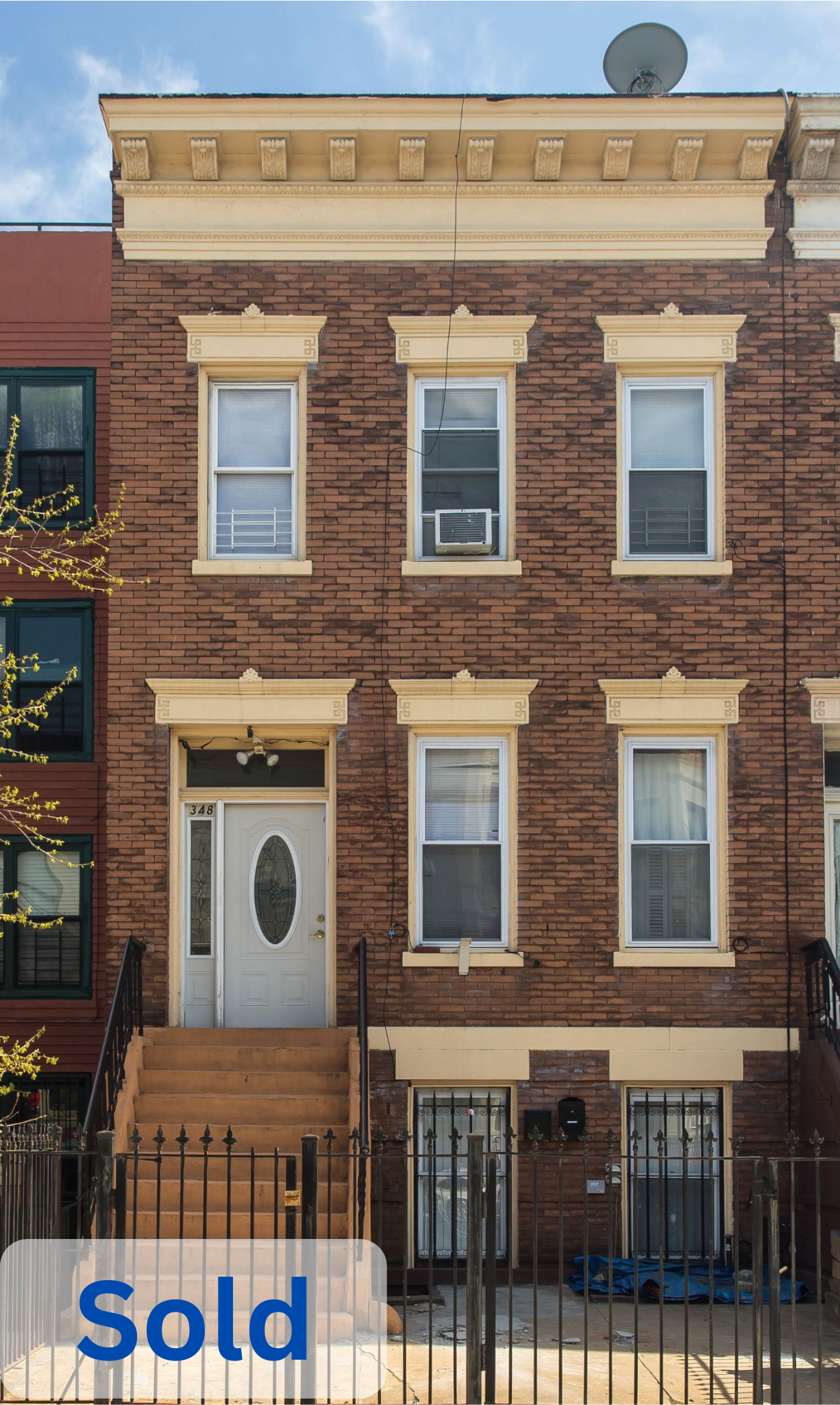 Three-story brick residential building with stairs leading to the front door, windows on each floor, and a surrounding metal fence. The door has the number 348 above it. The top floor has an air conditioning unit in the middle window, and there is a satellite dish on the roof. The bottom left corner has a blue and white 'sold' sign.