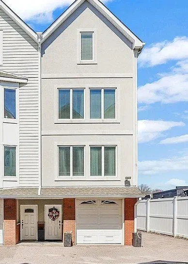 Three-story white house with brick base, garage, and front door decorated with a wreath, under a blue sky with clouds.