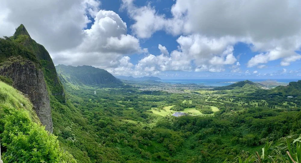 Lush green valley with mountains and clouds, overlooking the ocean in the distance.