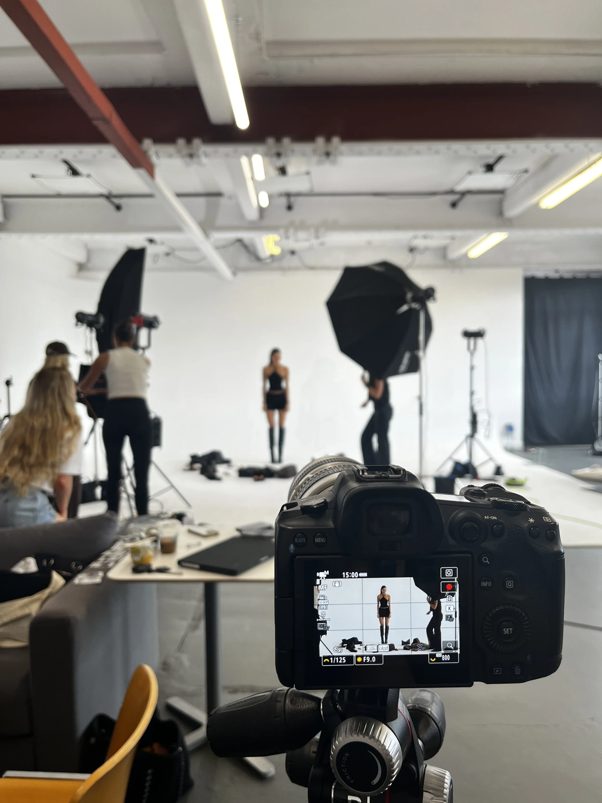 Photo of a photography studio set up with a model standing in front of a white backdrop, surrounded by lighting equipment and crew members. The camera on a tripod is focused on the model.