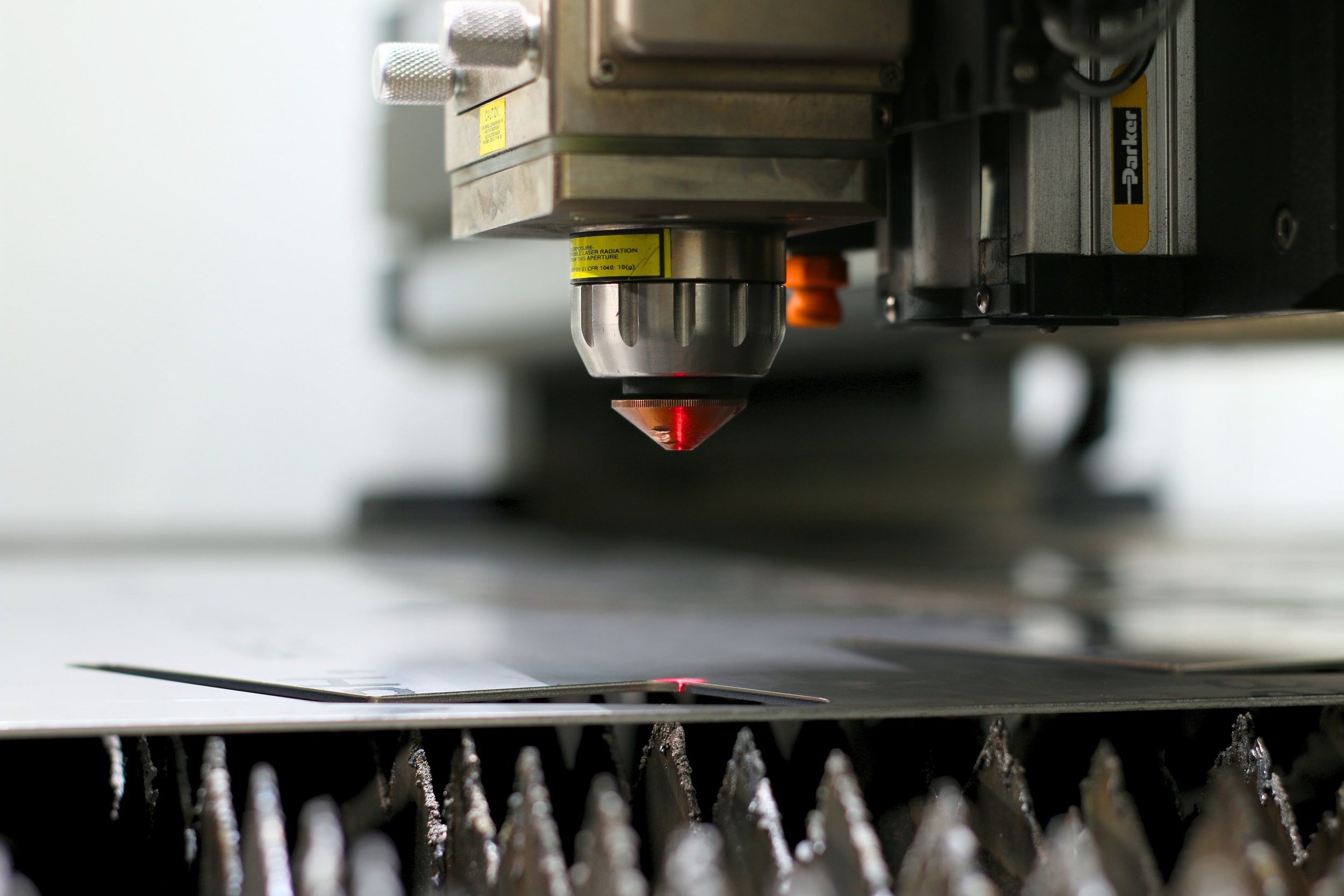 Close-up of a laser cutter machine in operation, with a red laser beam focused on a metal sheet, surrounded by metal components and industrial equipment.
