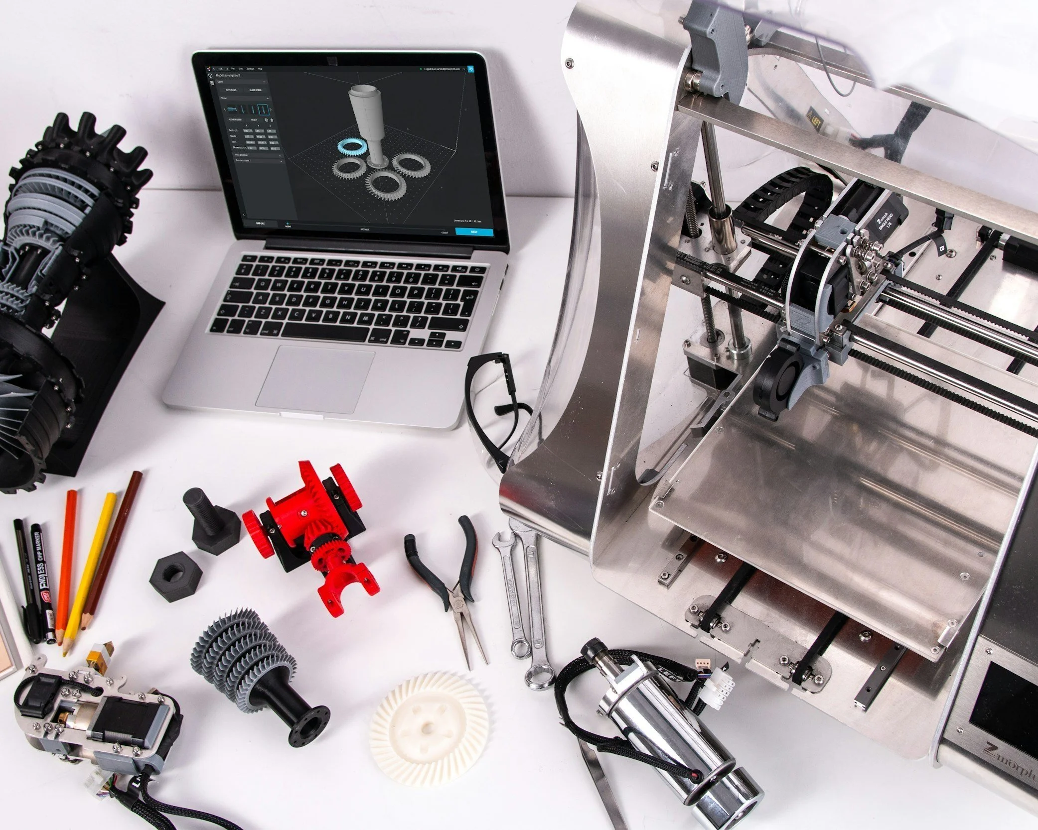 A workspace featuring a laptop displaying 3D modeling software, a robotic arm on the left, various mechanical parts and tools on the table, and a partially assembled mechanical device or 3D printer on the right.