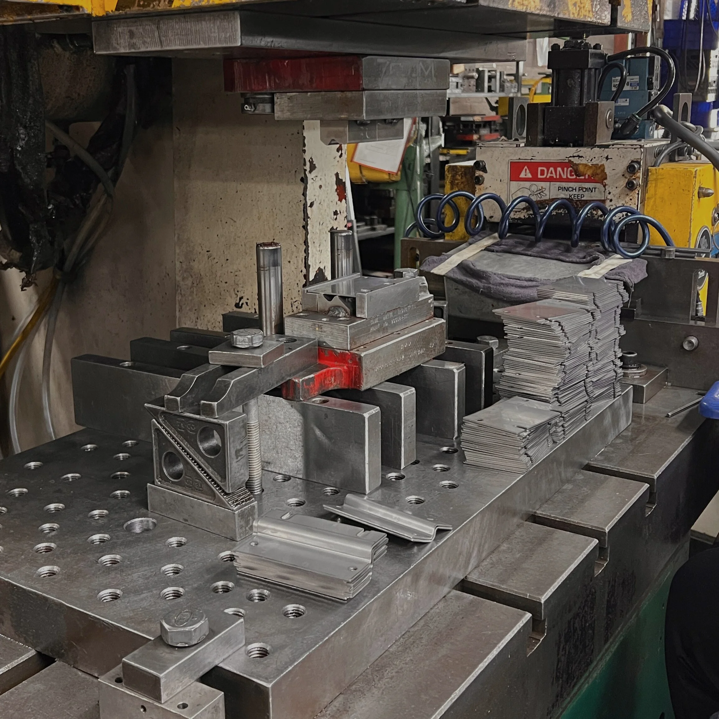 Metal stamping machine working with stacked metal pieces on the work surface, surrounded by industrial tools and equipment in a workshop.