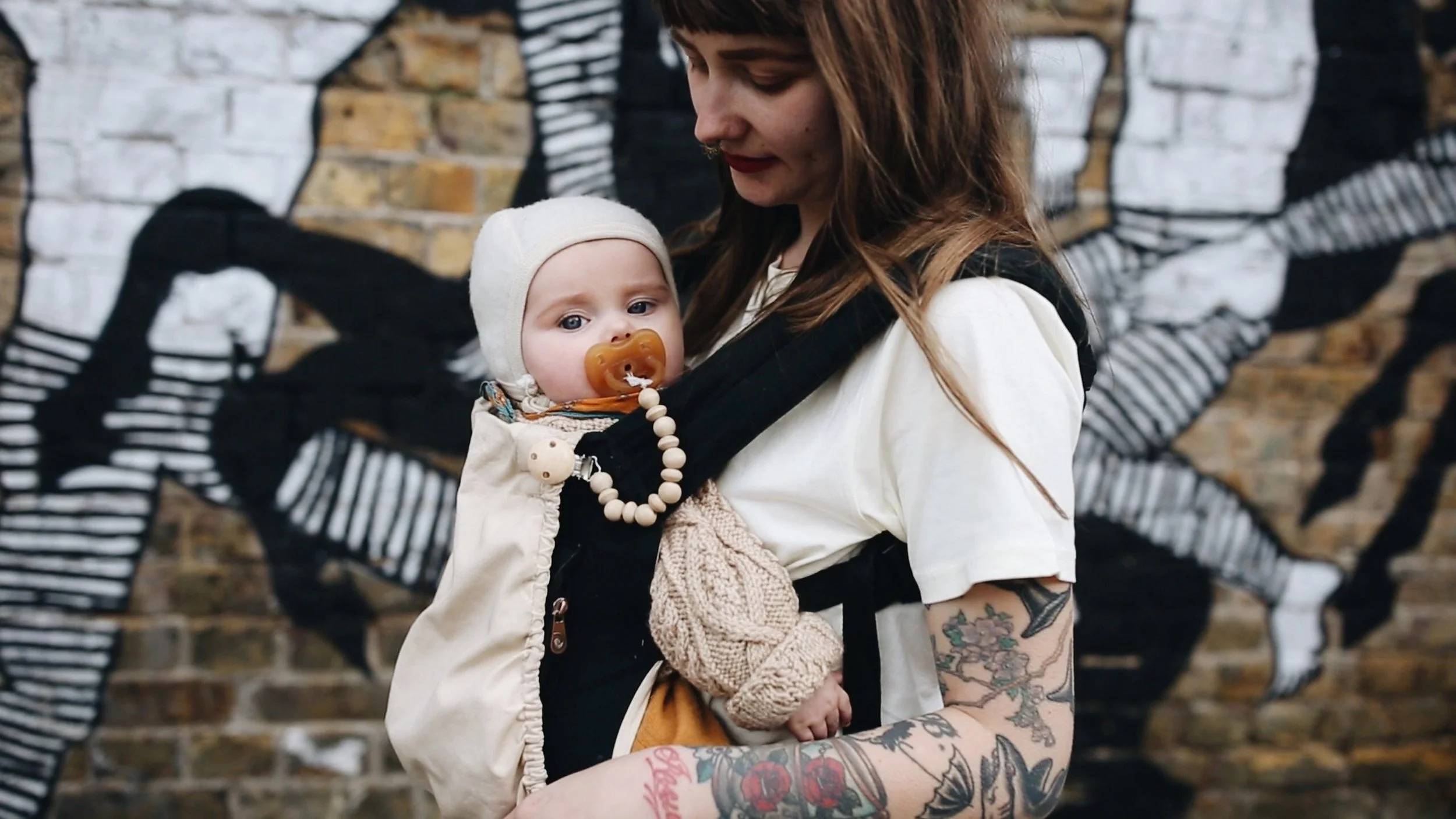 A woman holding a baby in a baby carrier. The baby has a pacifier and wears a hat. The woman has tattoos on her arm and is standing in front of a brick wall with a black and white mural.